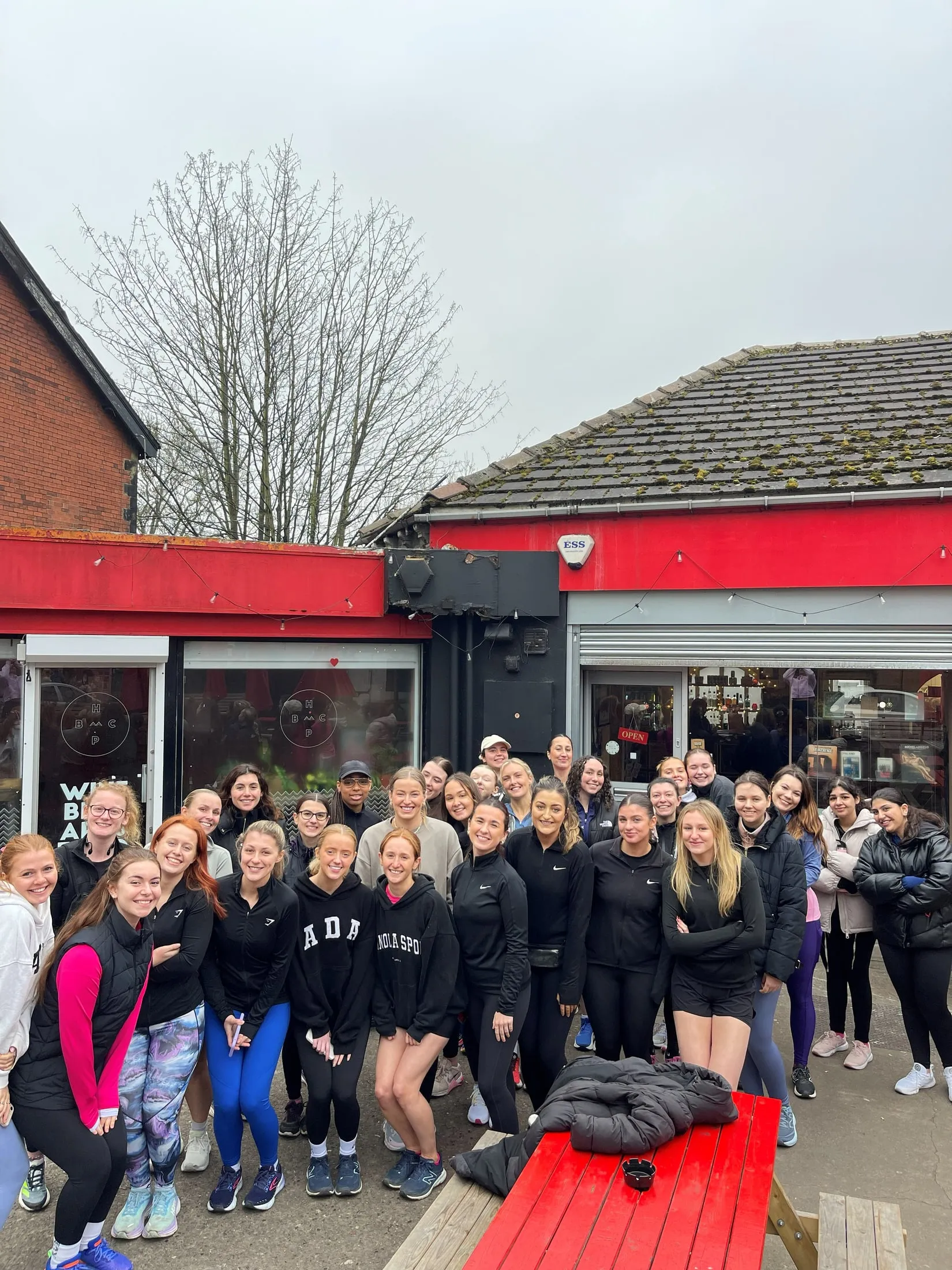 A big group in activewear posing outside a red-fronted building under a grey sky