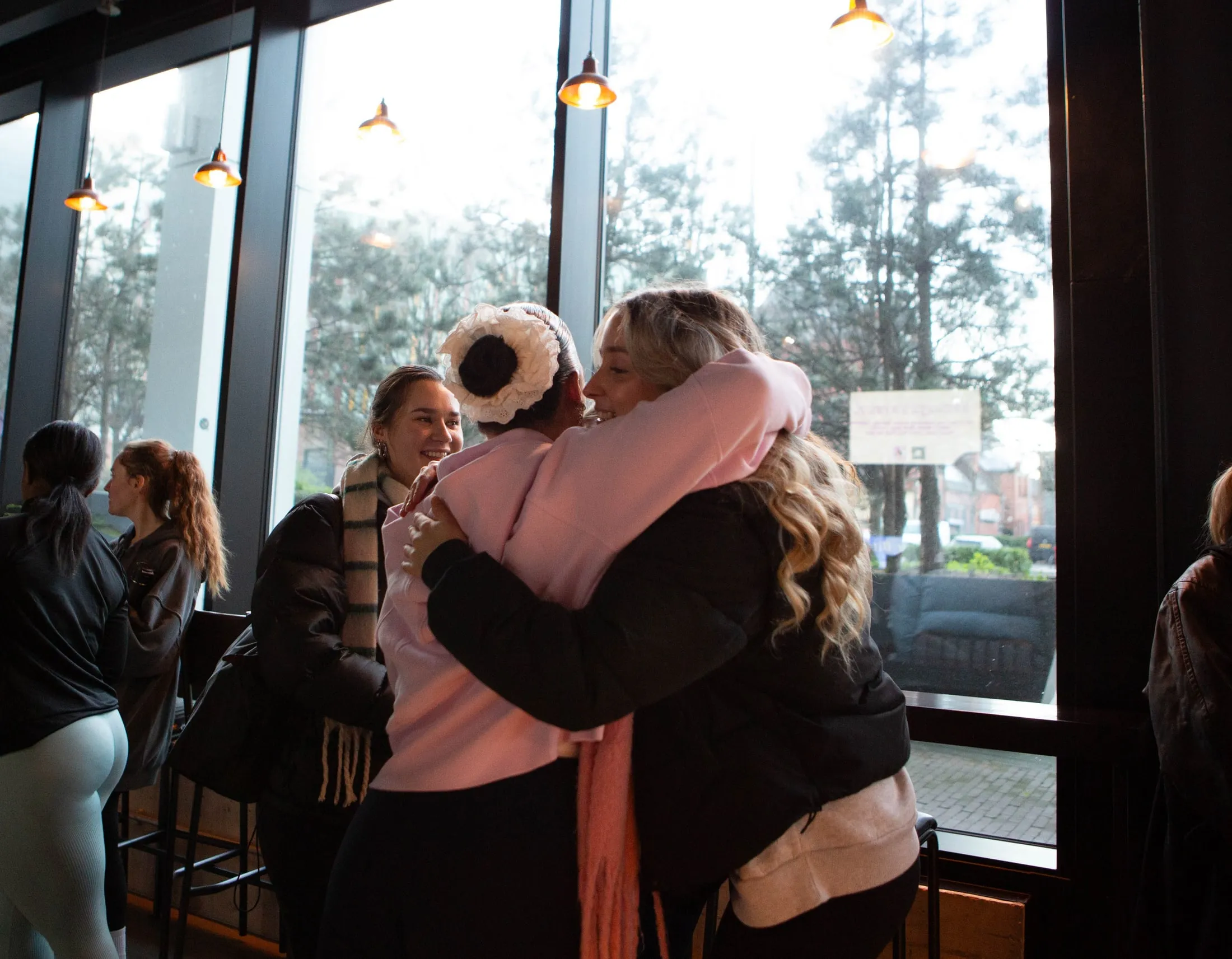 Two women sharing a big hug by tall windows, with others smiling and chatting behind