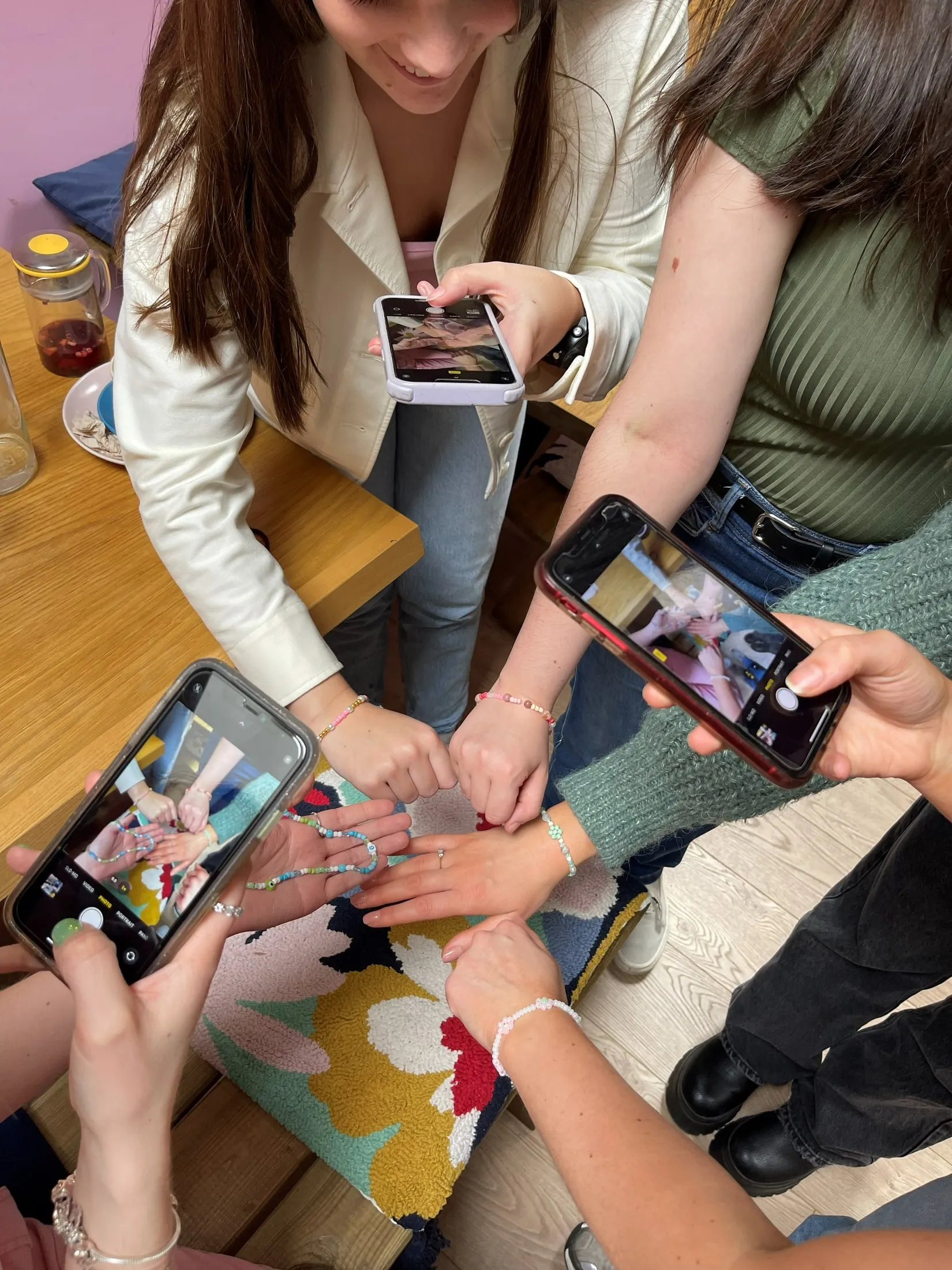 Hands showing off beaded bracelets over a flowery cushion while three phones take photos