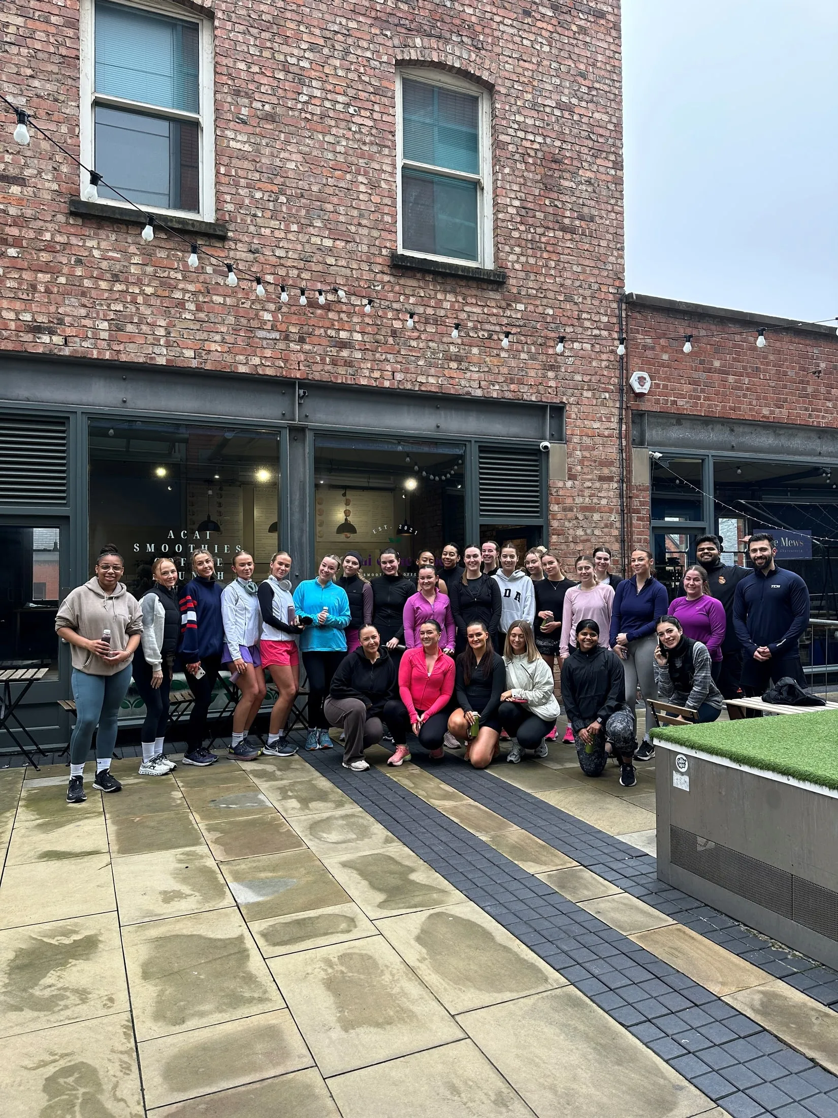 A big group in activewear posing outside a red-brick building hung with festoon lights