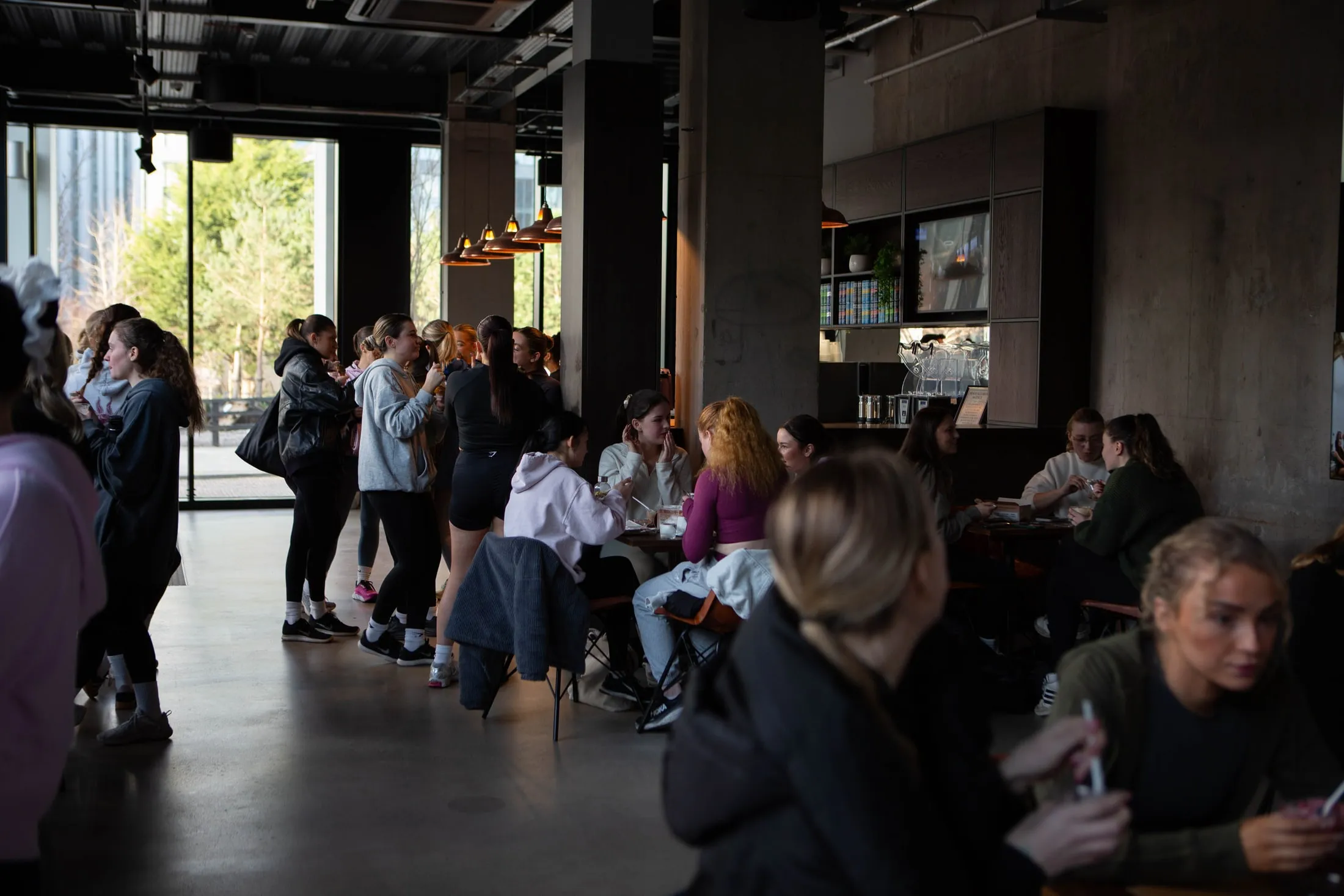A café full of women in gym wear, some standing by tall windows, some eating at tables
