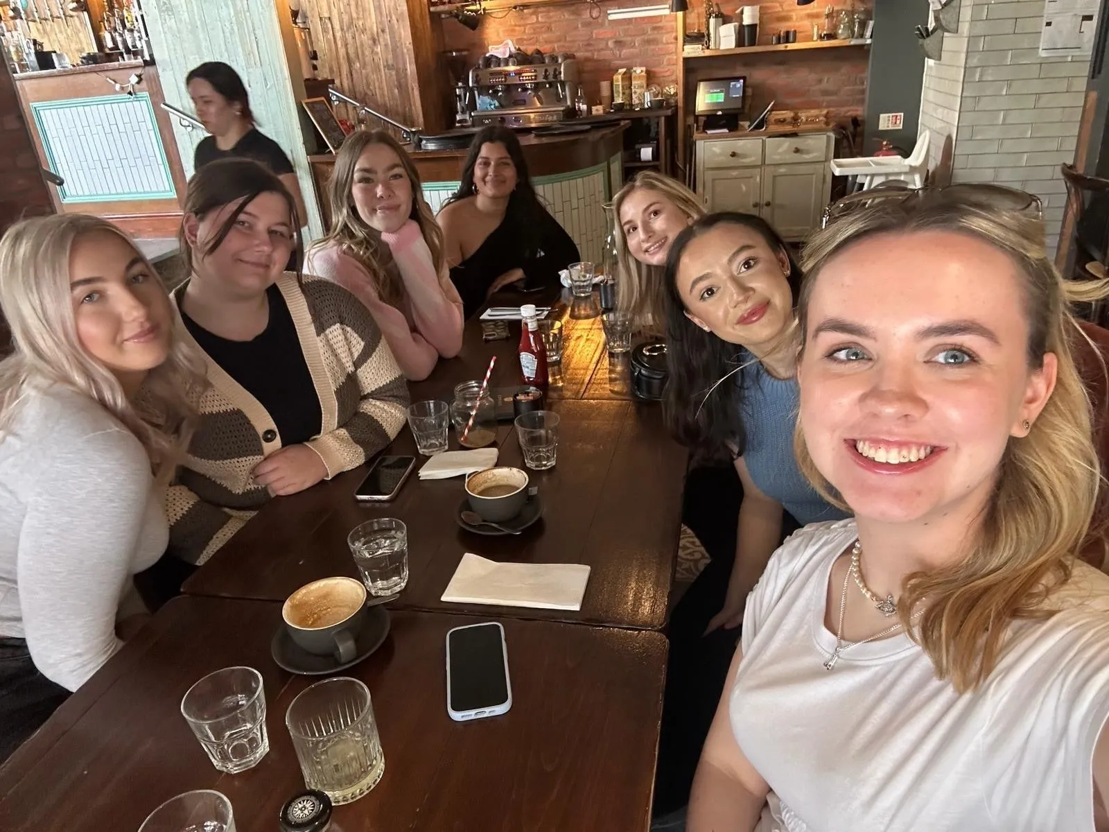 A selfie of seven women round a café table with coffees and glasses of water