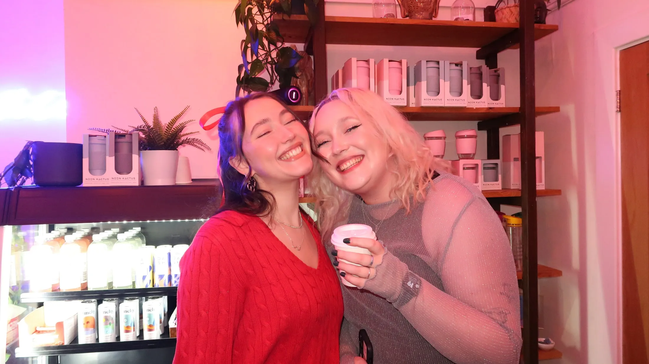 Two women cheek to cheek, beaming, by shelves of boxed cups and a drinks fridge in pink light