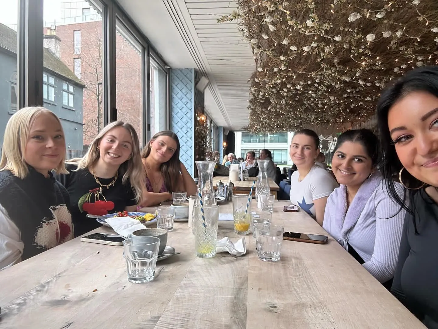 Six women round a long table under a ceiling of white flowers and fairy lights