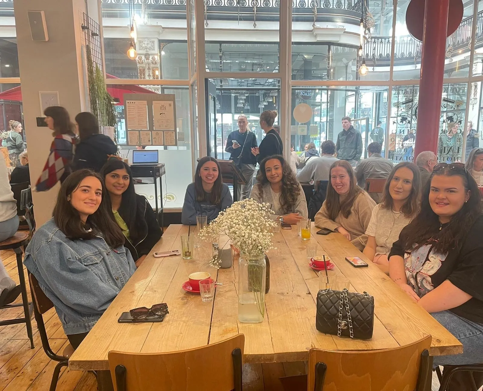 Seven women round a long café table with a vase of white flowers and red coffee cups