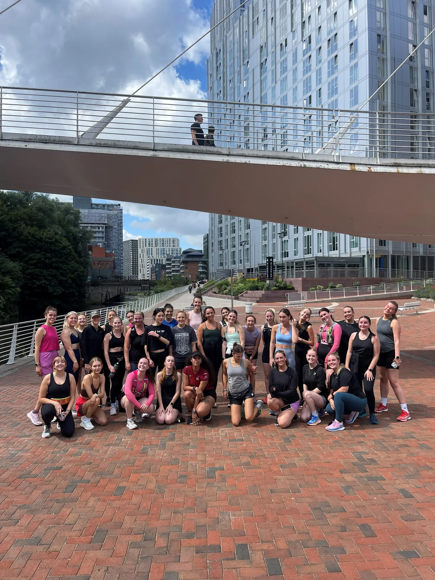 A big group in activewear posing on a brick walkway beneath a footbridge on a sunny day