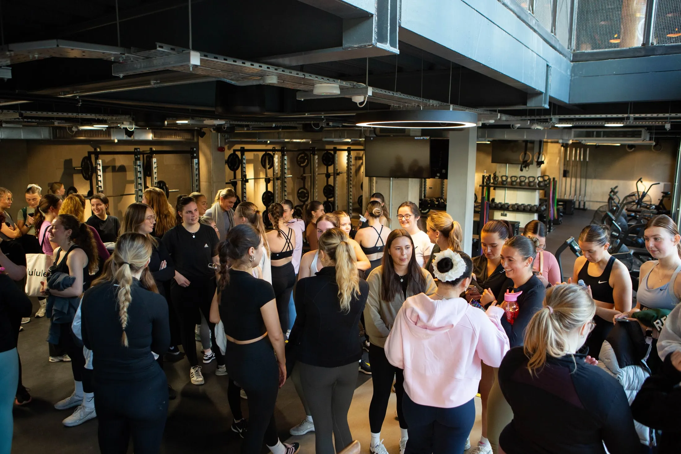 A big group of women standing in a loose circle in a gym, with weight racks and bikes behind