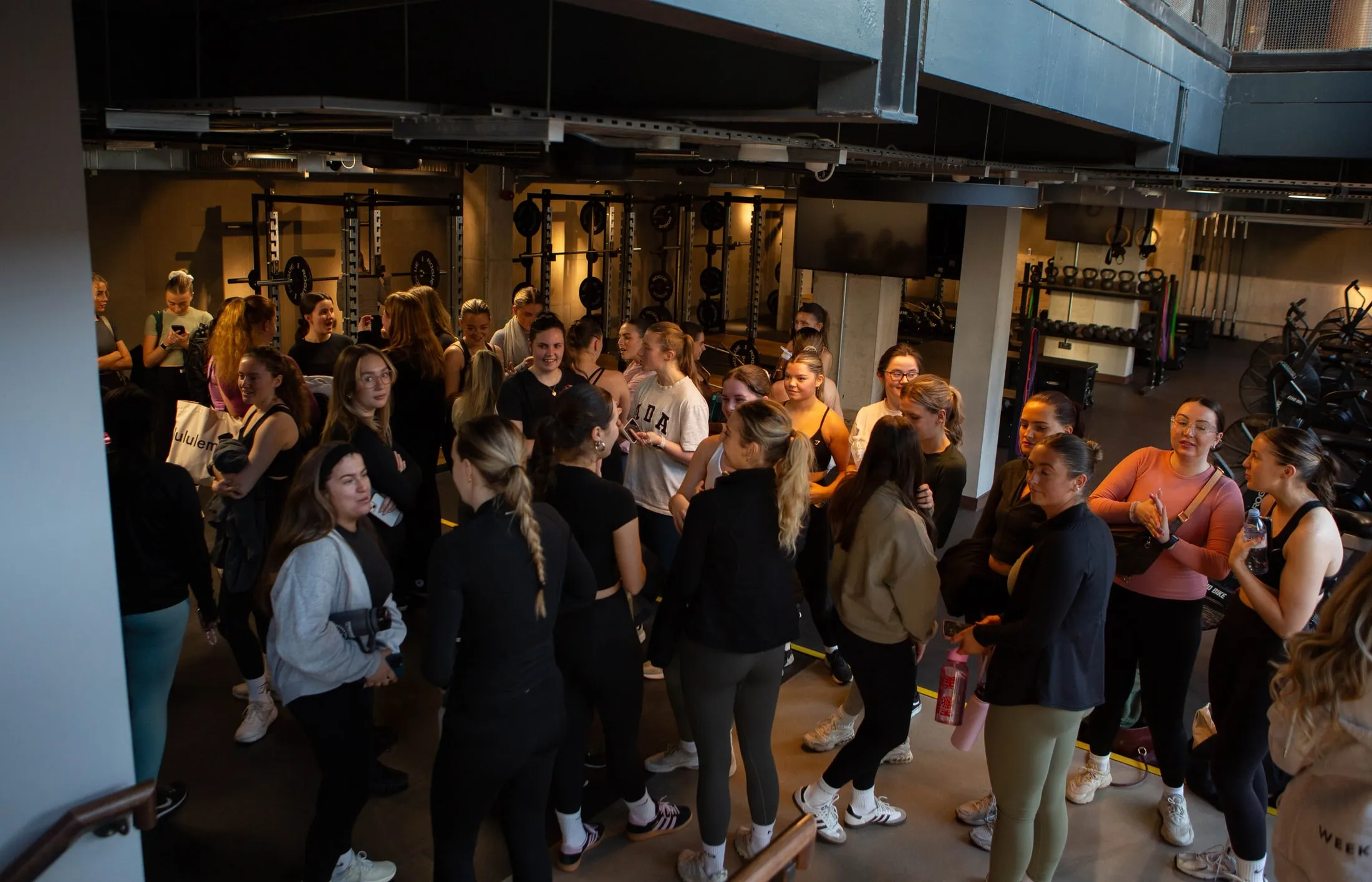 A big group of women in activewear chatting in a gym with squat racks, seen from above