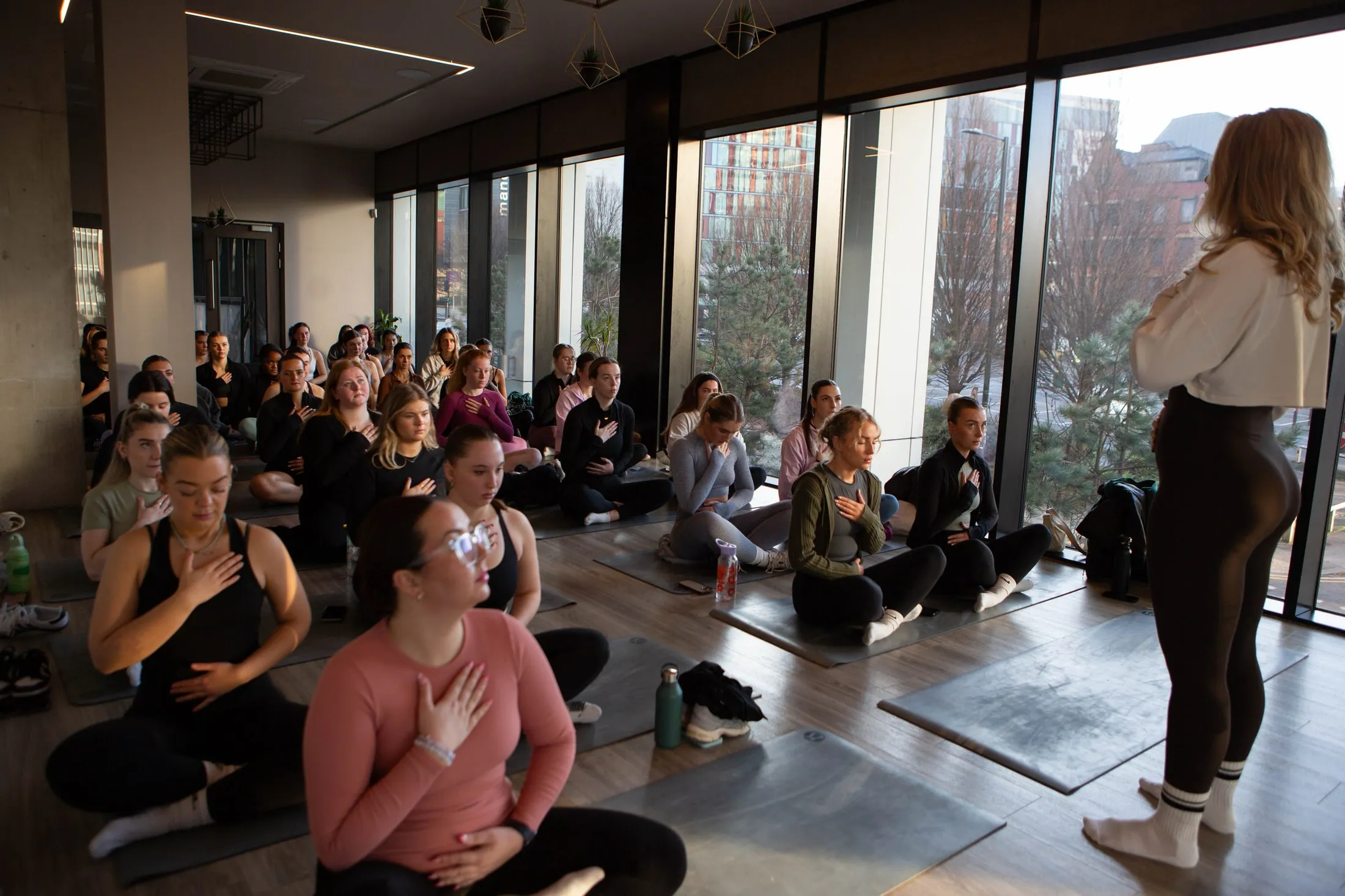Women sitting cross-legged on mats by tall windows, hands on hearts, as a woman stands facing them