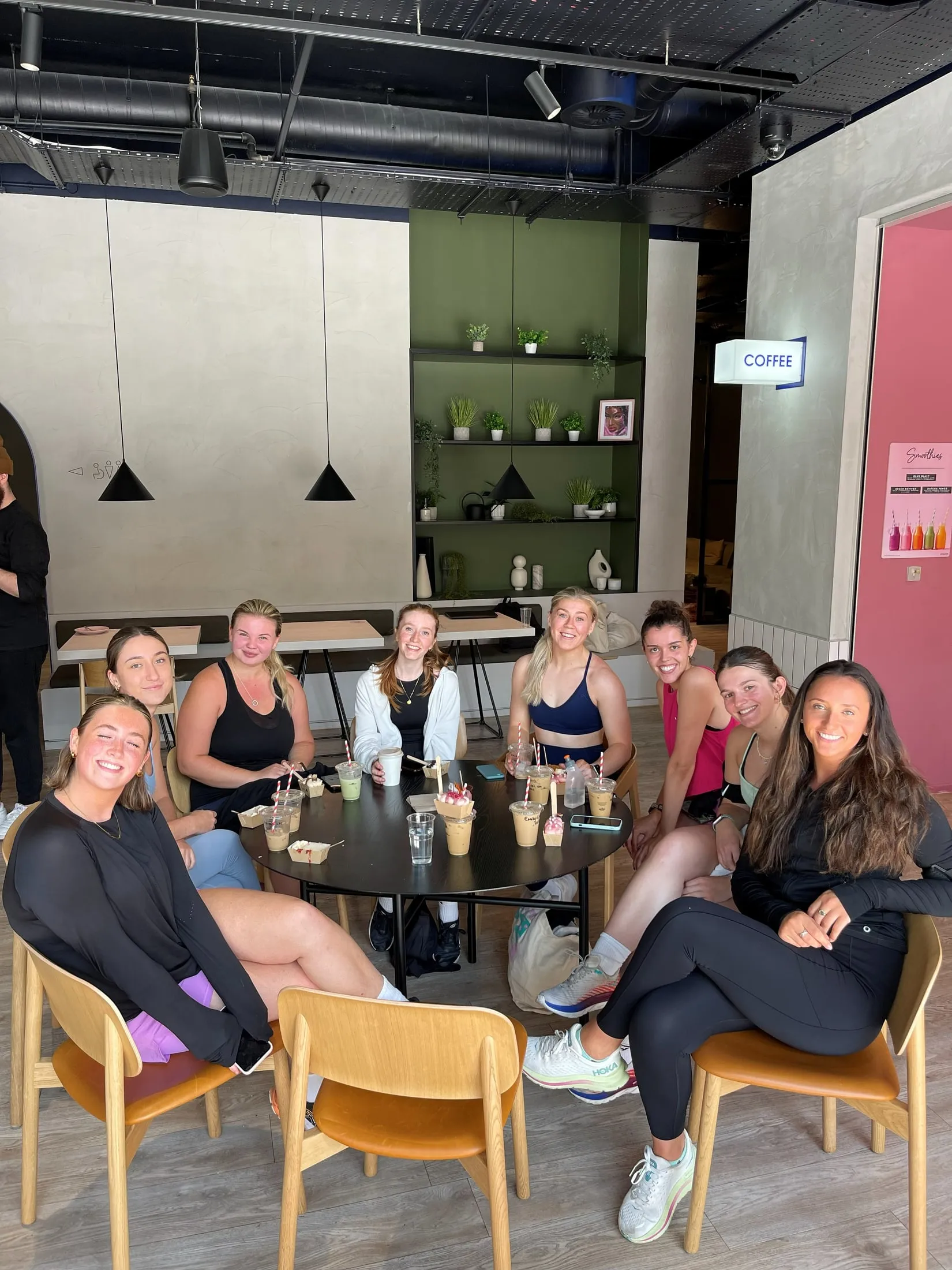 Eight women in gym wear with iced drinks and snacks around a table in a café with green shelves