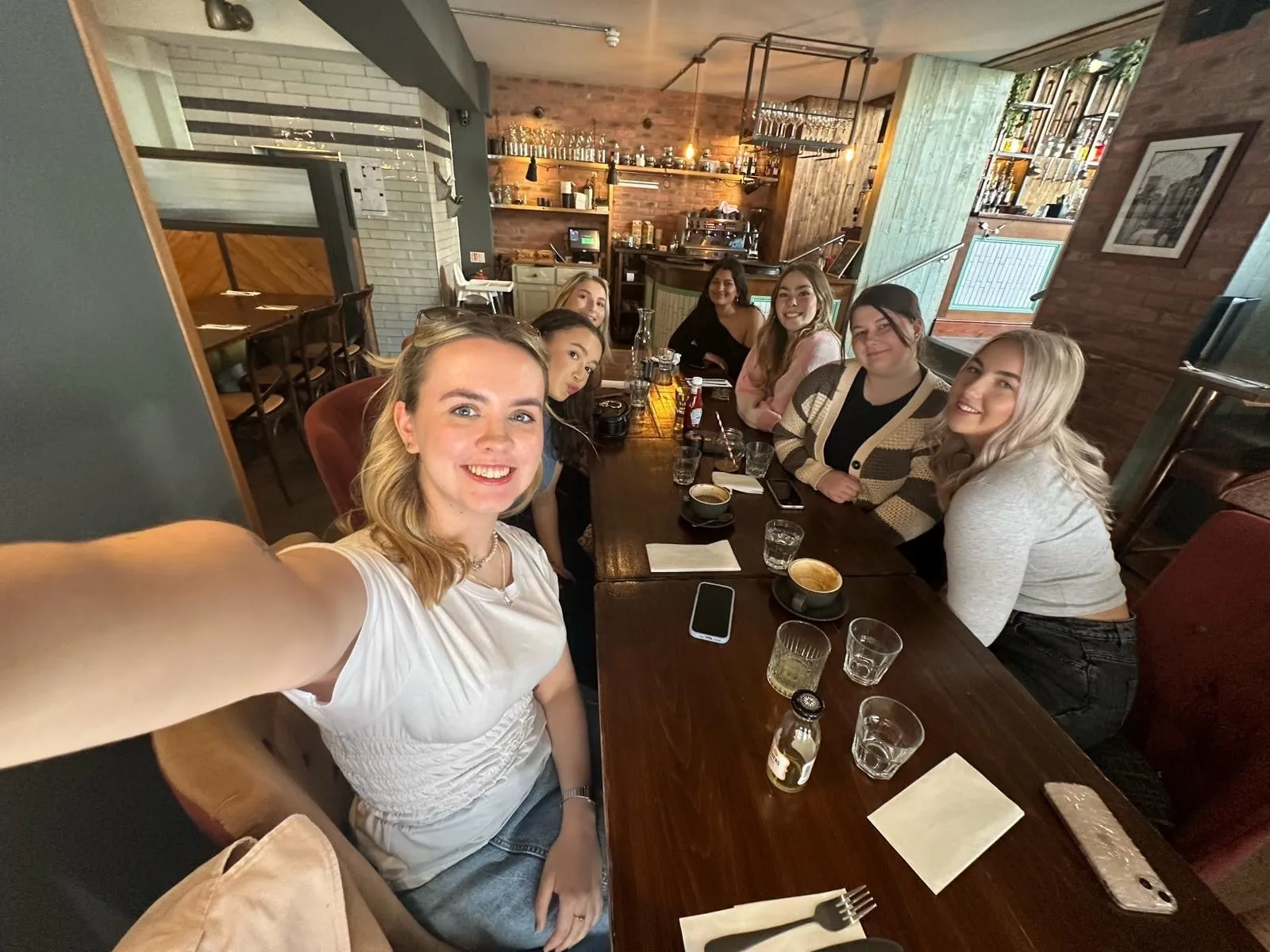 A wide-angle selfie of seven women round a long table, with a brick-walled bar behind