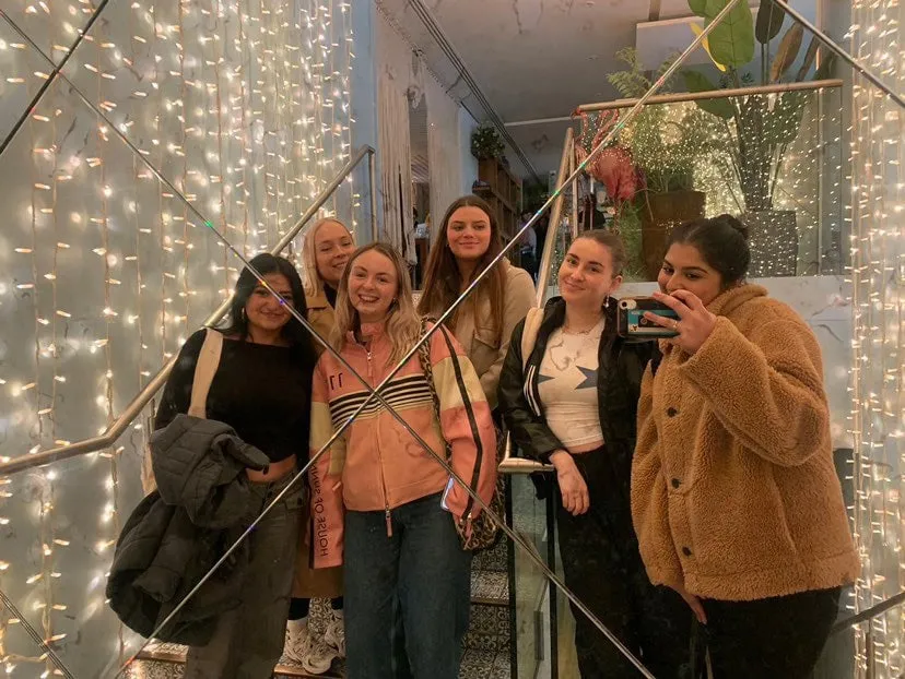 Six women posing for a mirror selfie on a staircase framed by curtains of fairy lights