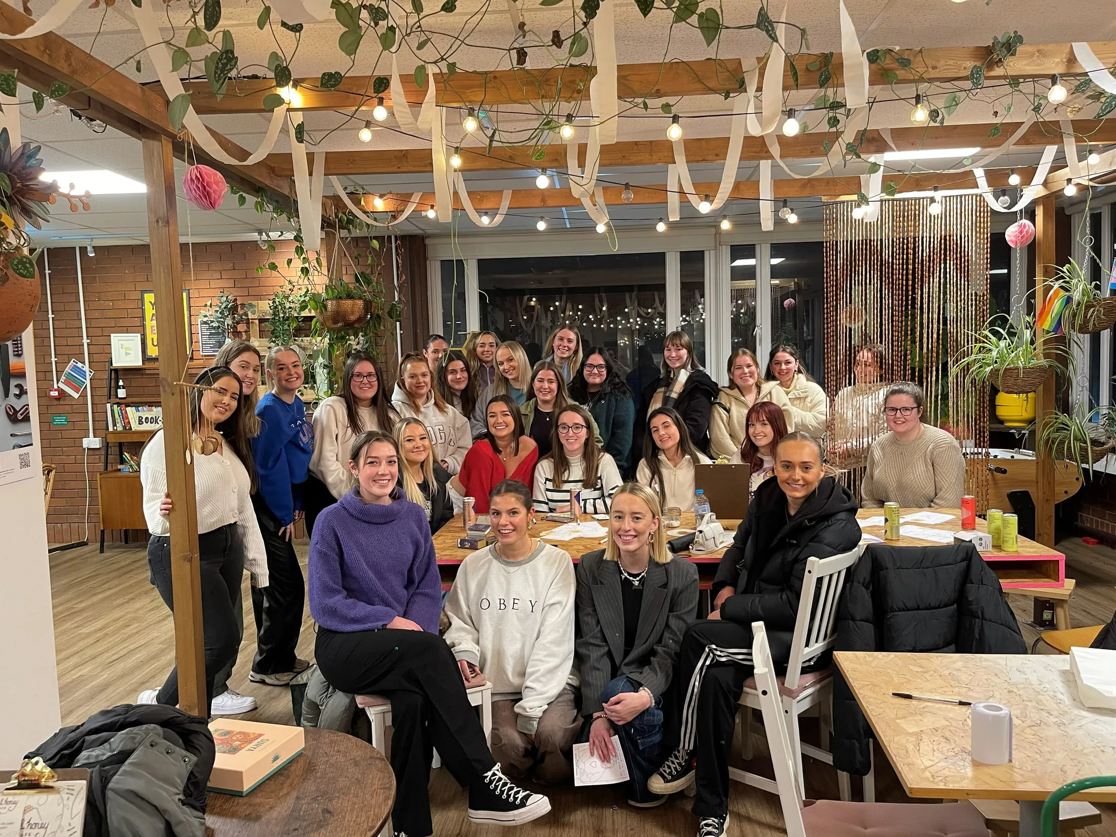A big group smiling around tables under a pergola hung with lights, plants and paper streamers