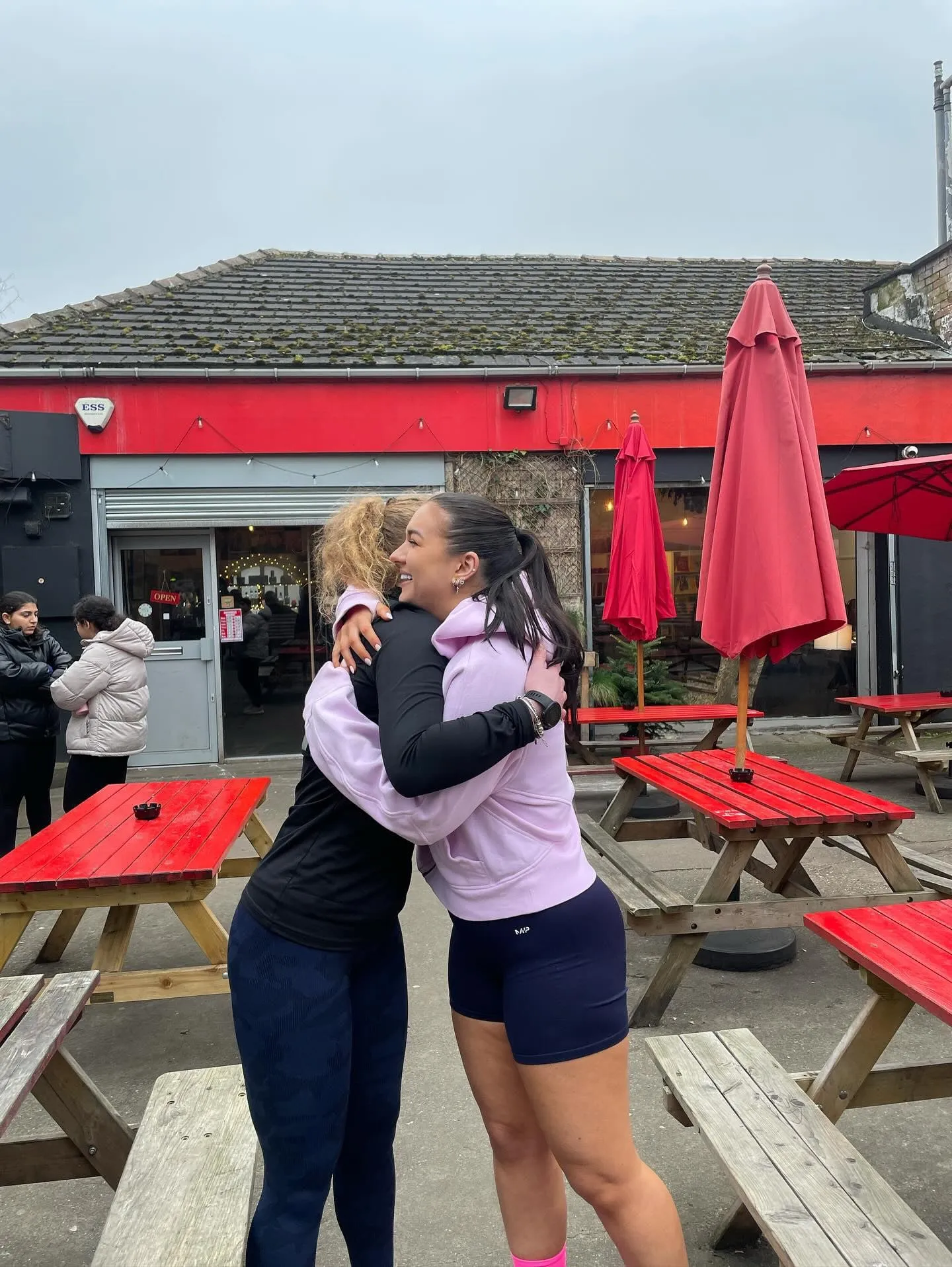 Two women in activewear hugging among red picnic tables and parasols