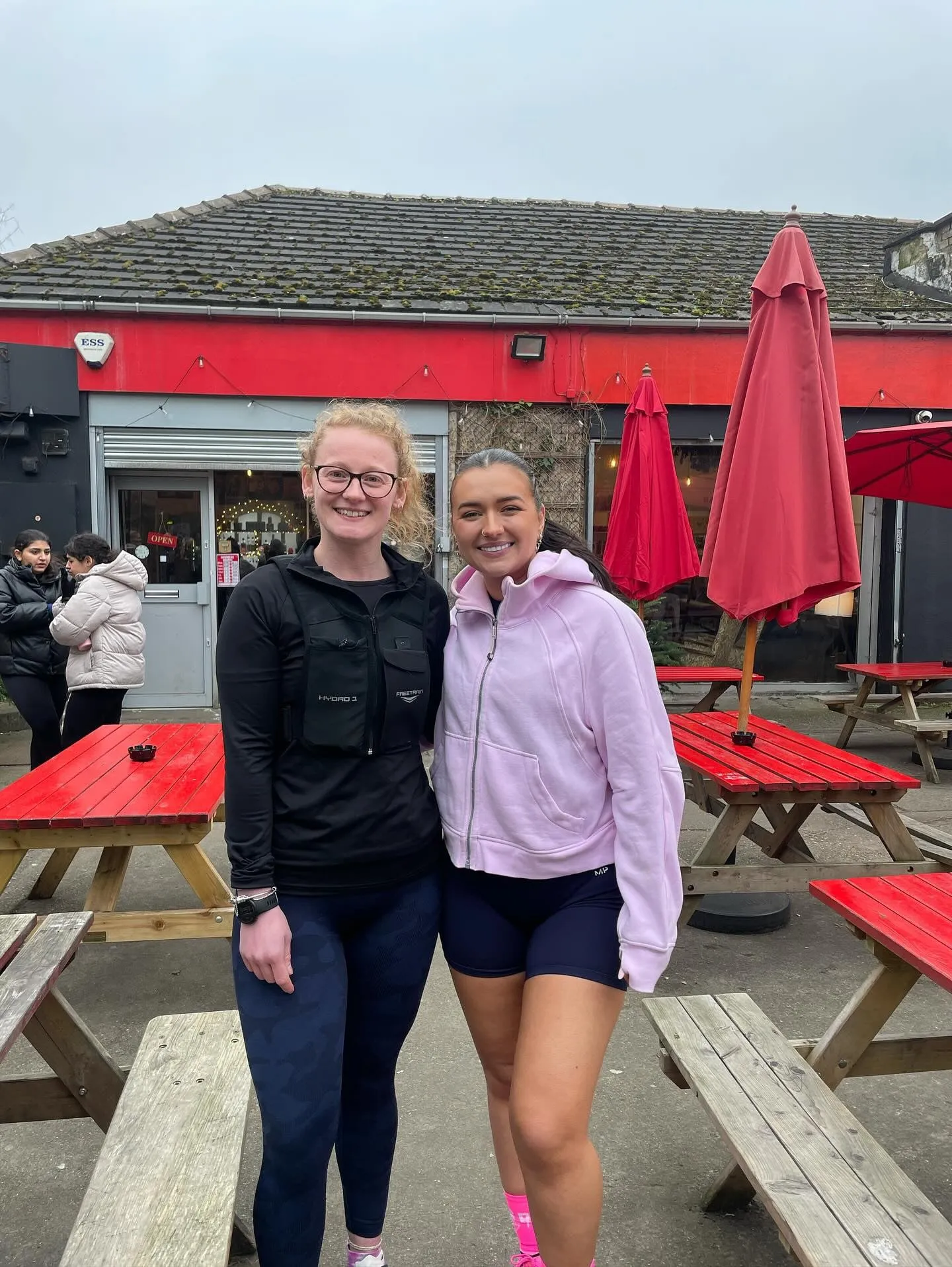 Two women in activewear smiling side by side among red picnic tables and parasols