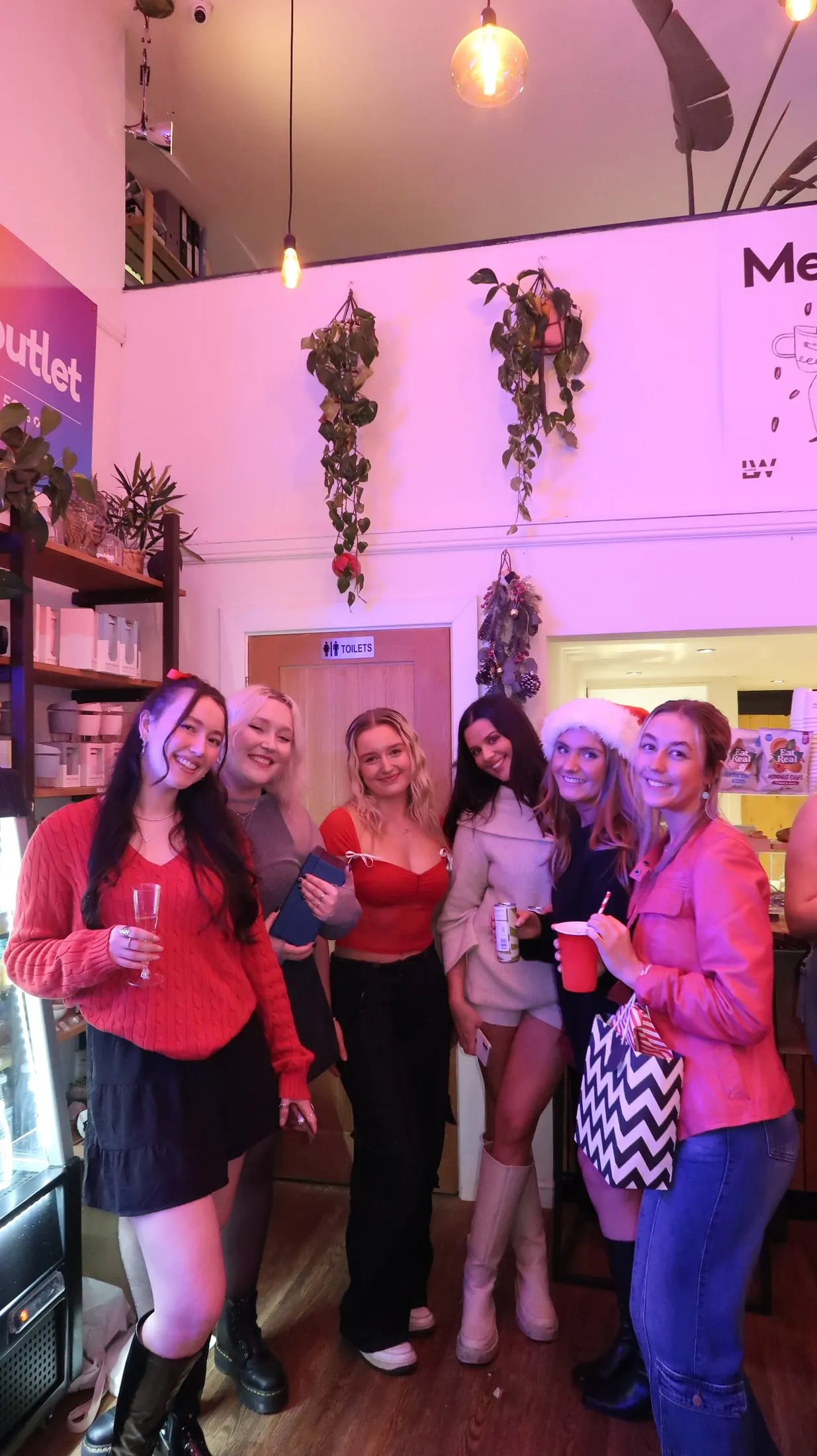 Six women posing under hanging plants in a pink-lit room, one in a Santa hat