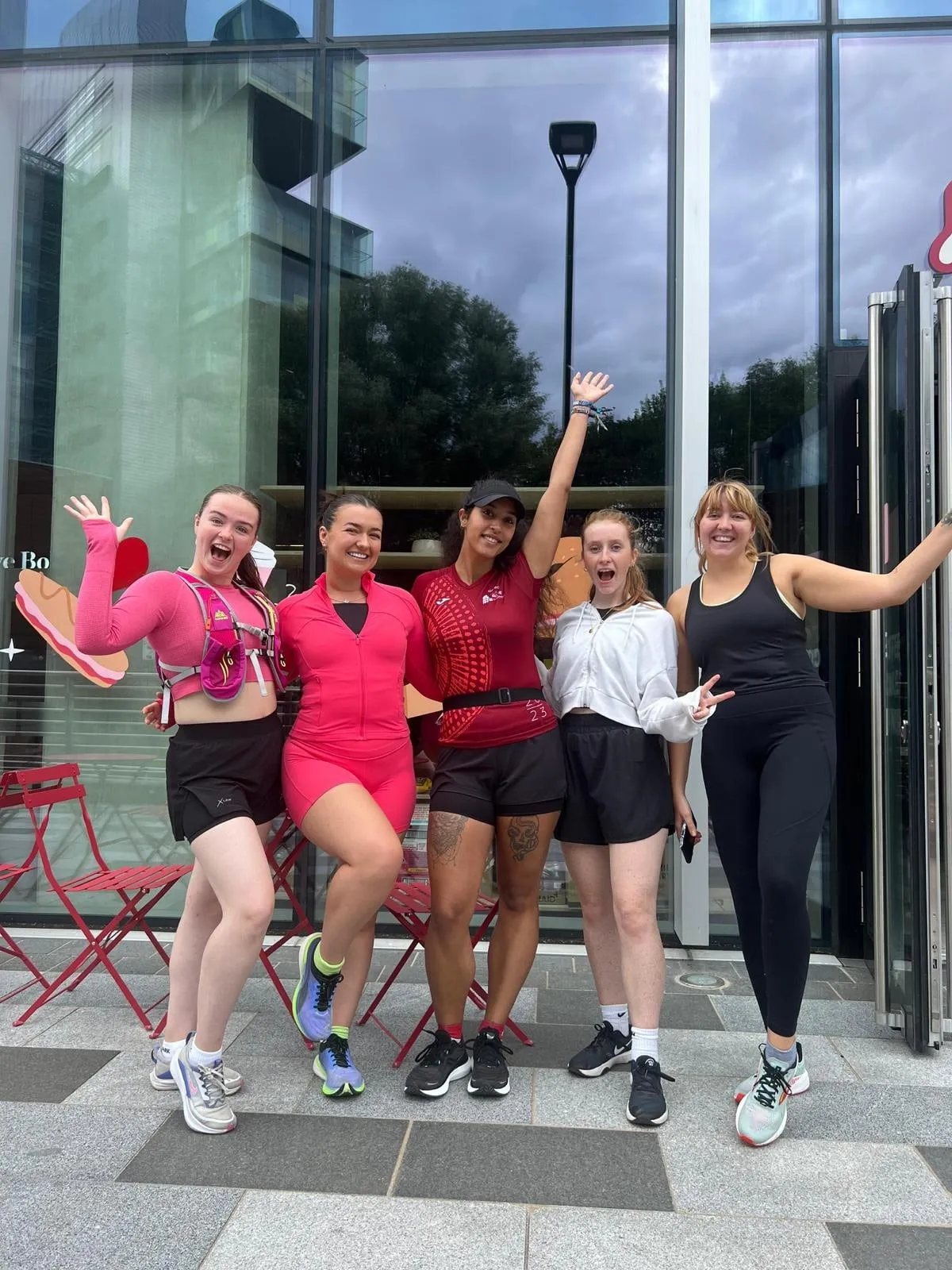 Five women in activewear striking playful poses outside a glass-fronted building