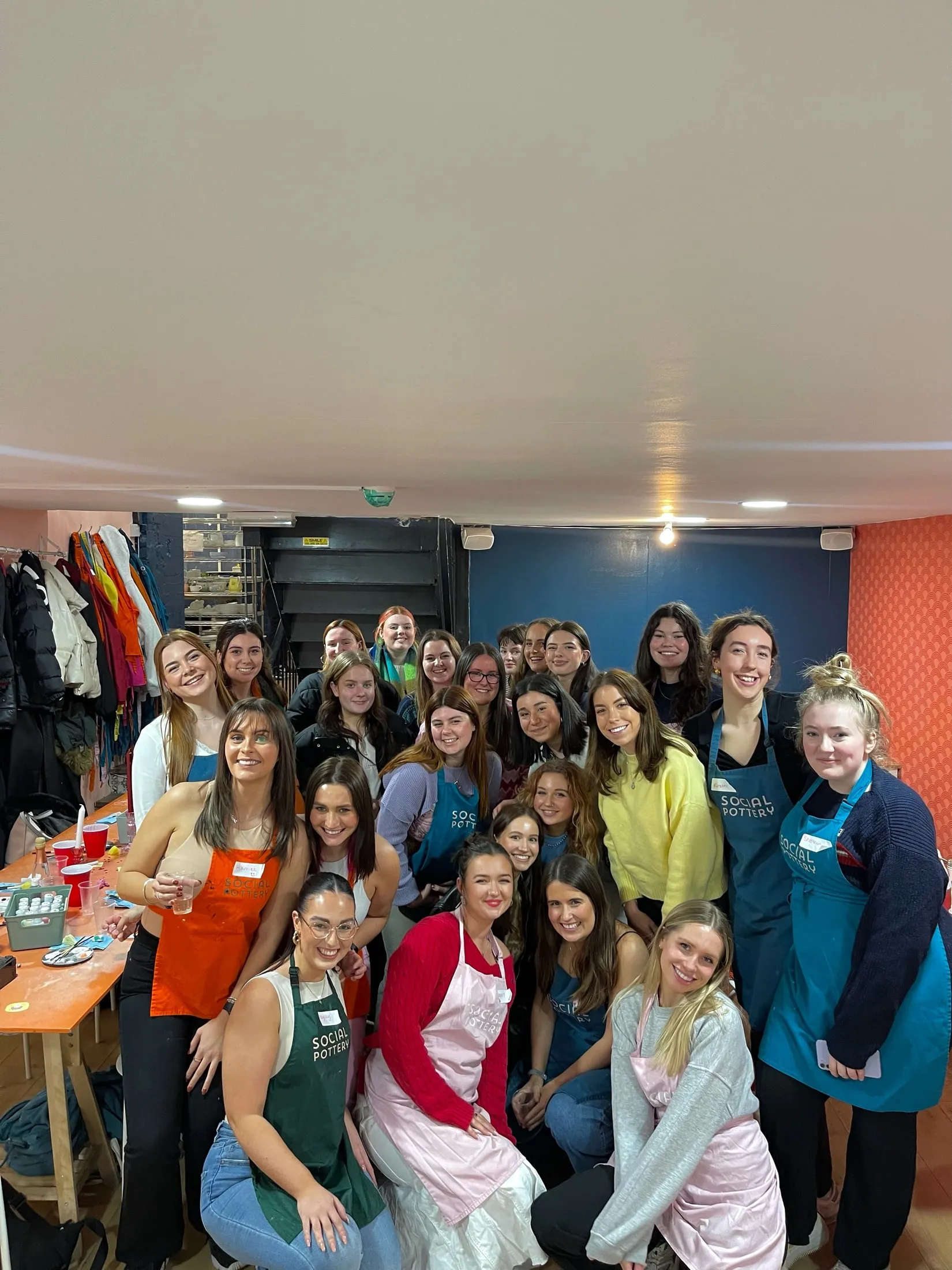A big group of women, many in Social Pottery aprons, posing together by a blue wall