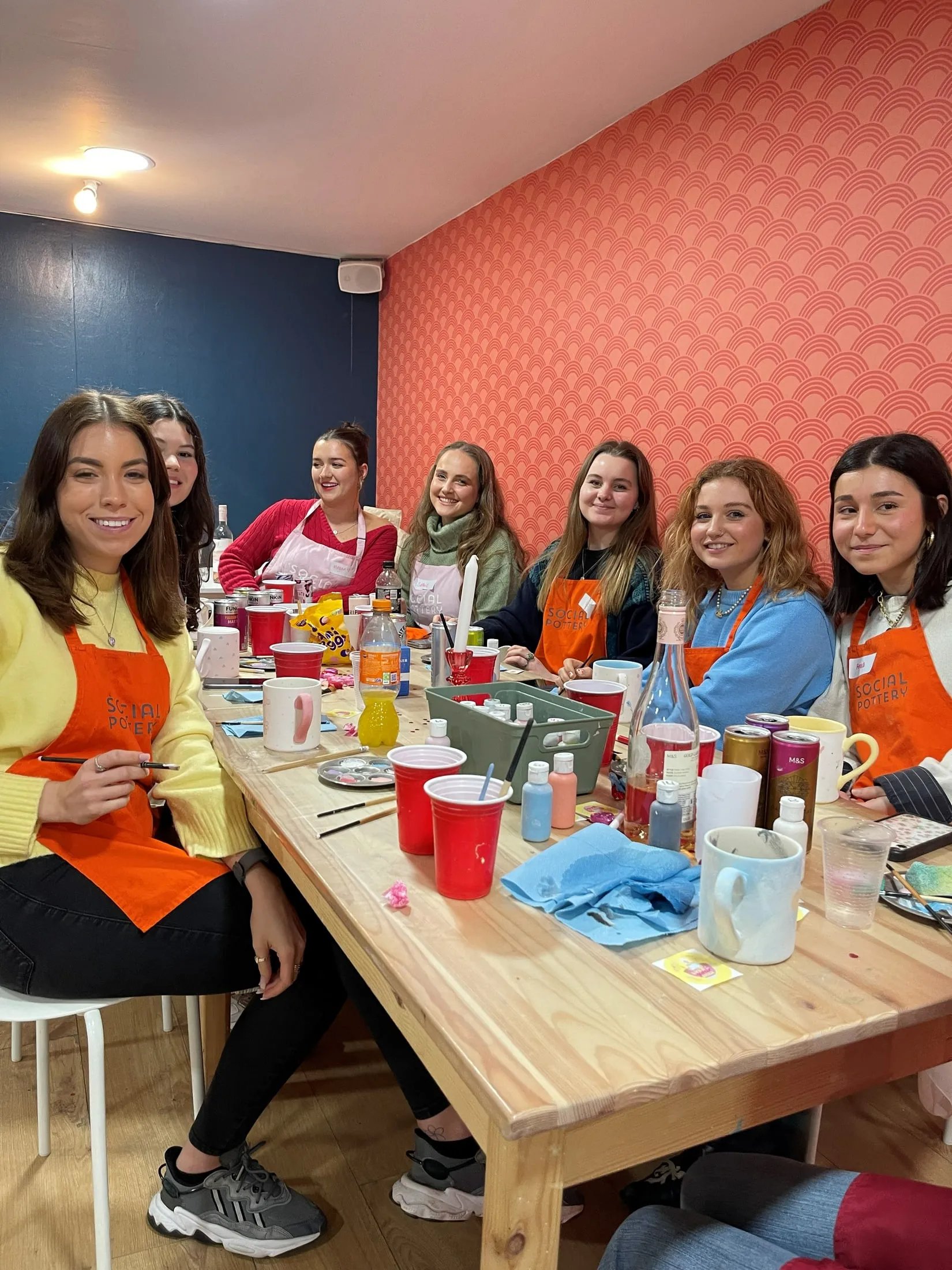 Women in orange and pink aprons painting mugs around a table by a coral patterned wall