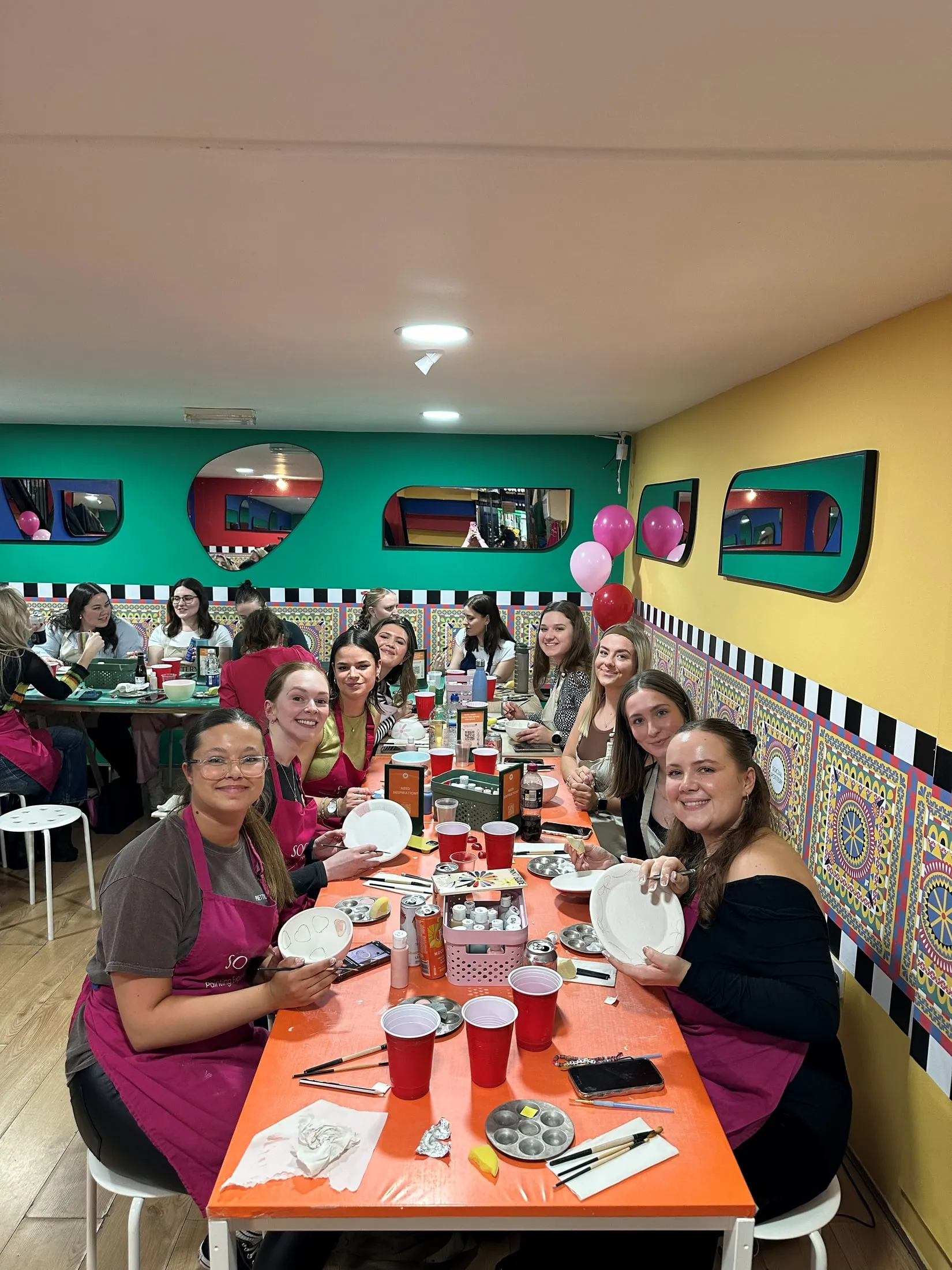 Women in pink aprons smiling as they paint plates and bowls along a bright orange table