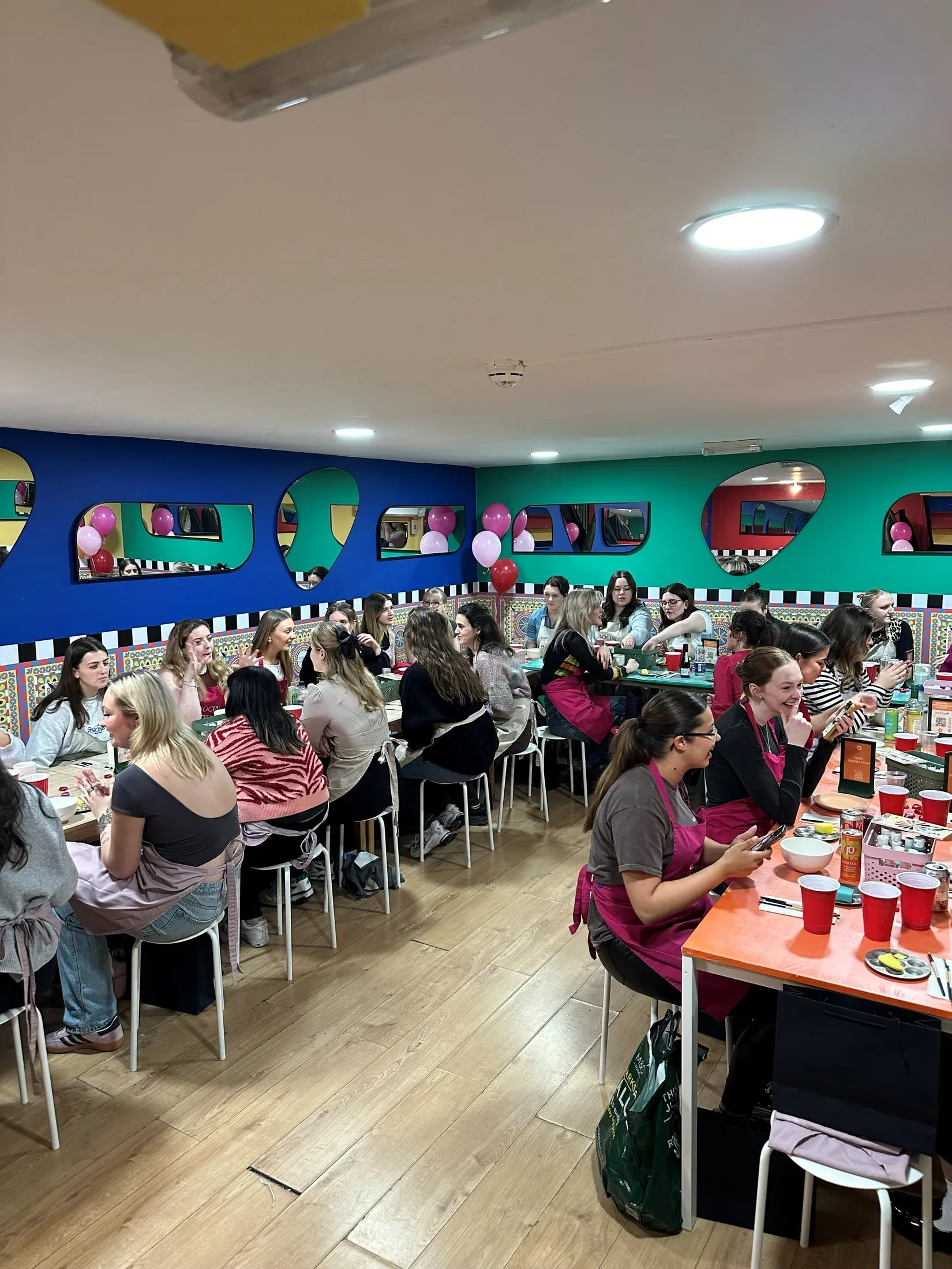 A big group in aprons chatting at long tables in a blue and green room with pink and red balloons