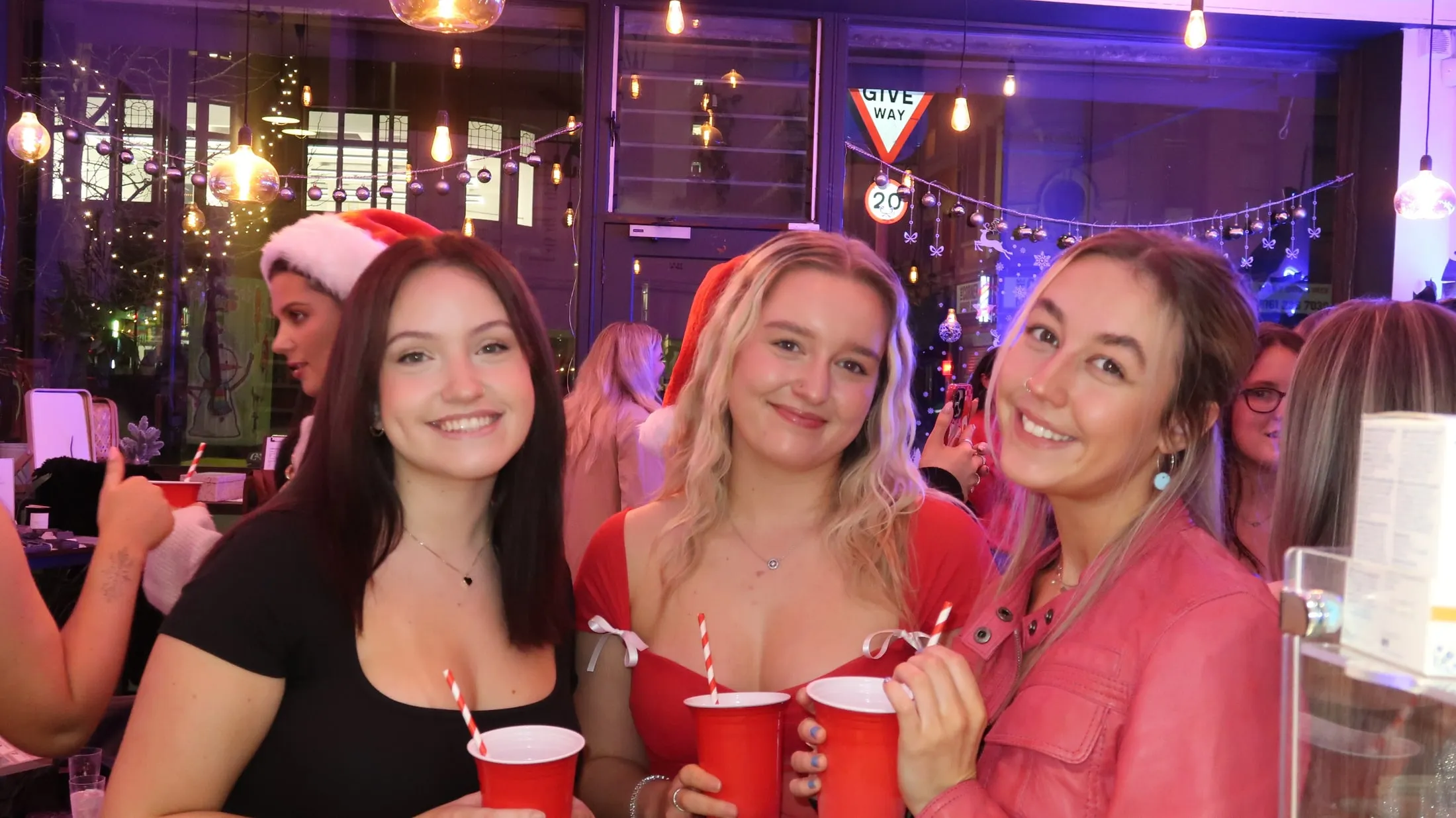 Three smiling women with red cups and candy-striped straws, baubles and string lights behind