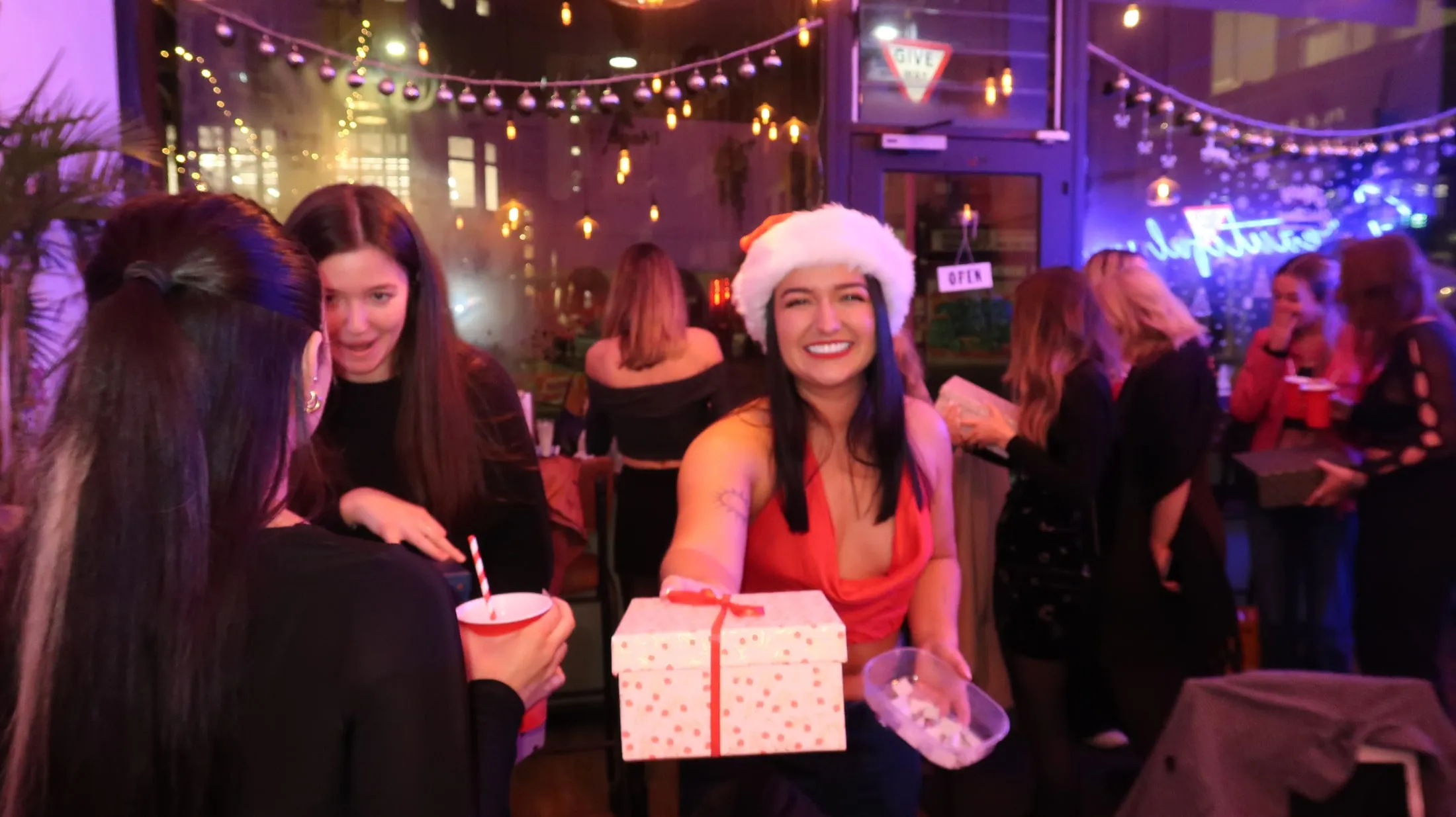 A woman in a Santa hat holding out a wrapped gift under strings of baubles and fairy lights
