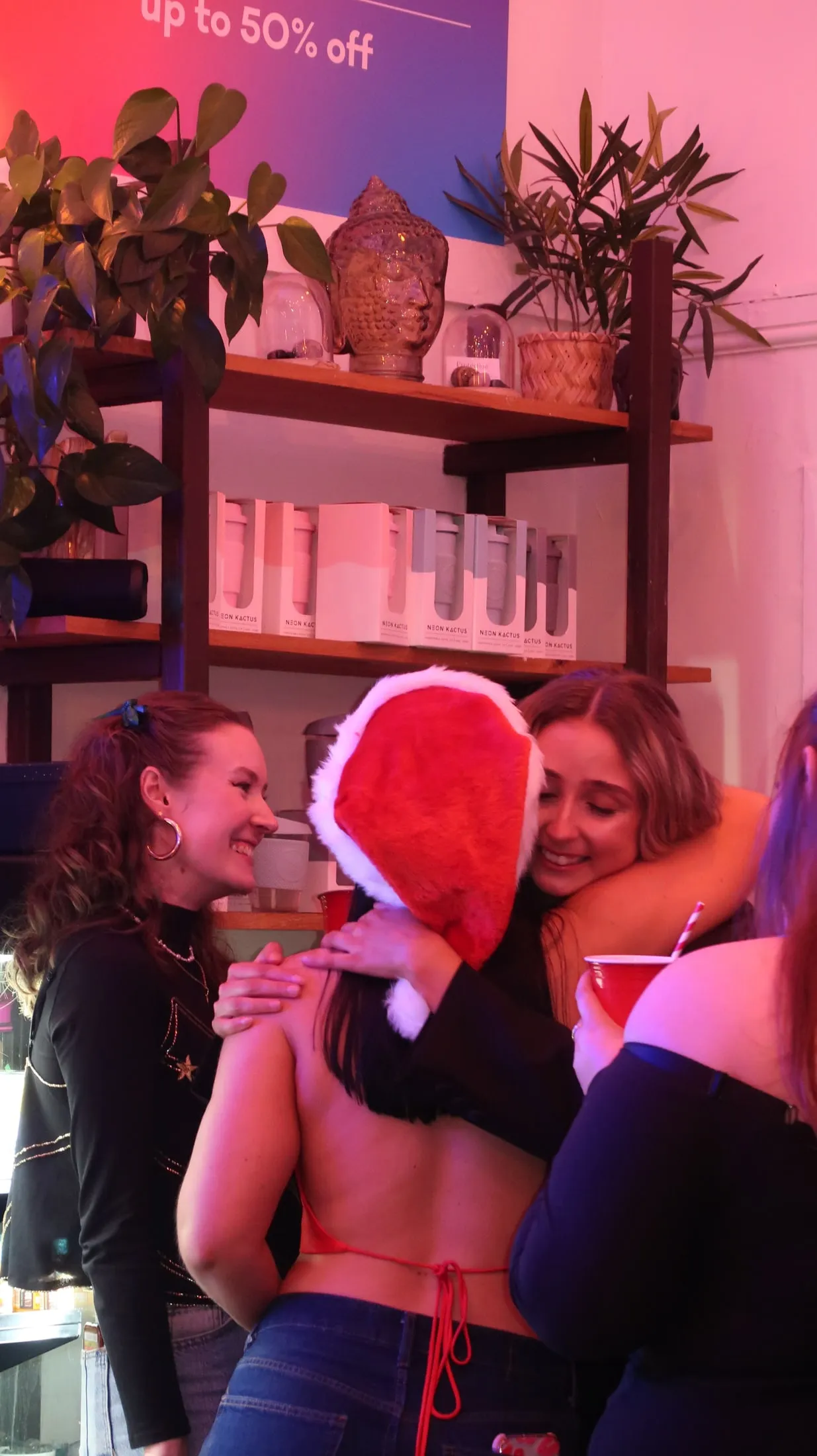 Two women hugging, one in a Santa hat, as another woman smiles beside them in a pink-lit room
