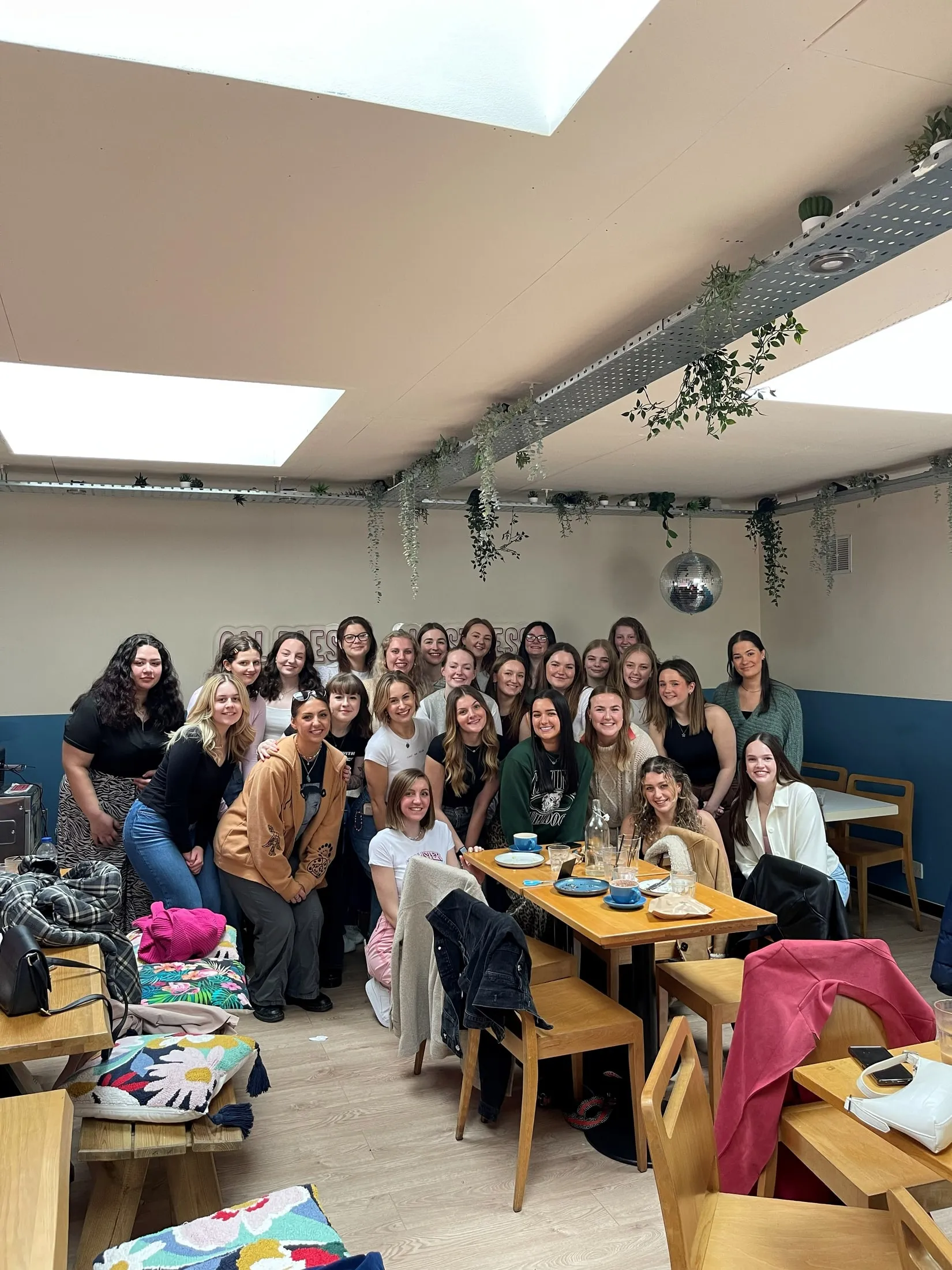 A big group of women posing in a room with skylights, hanging plants and a disco ball
