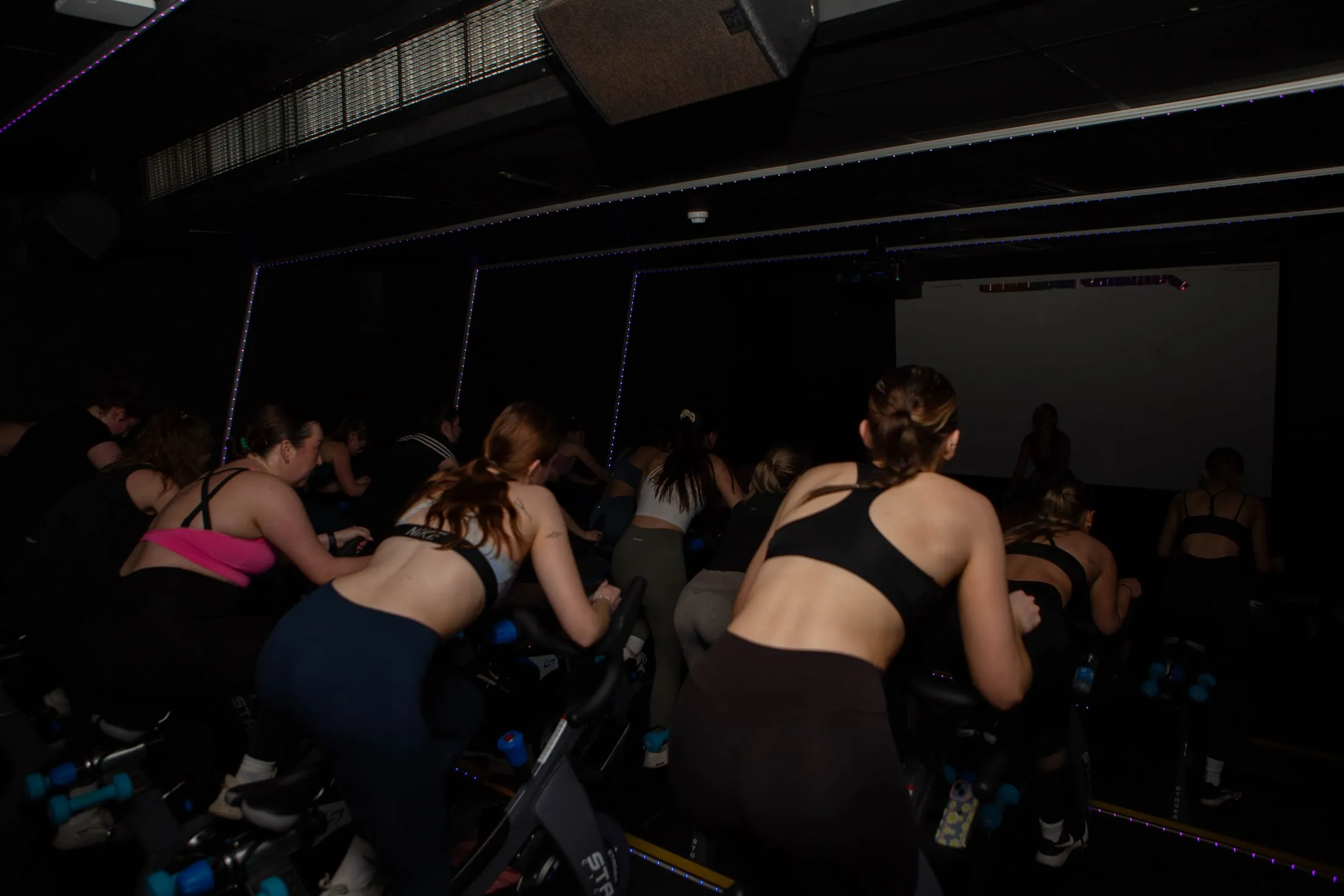 Women riding spin bikes towards a big lit screen in a dark studio, seen from behind