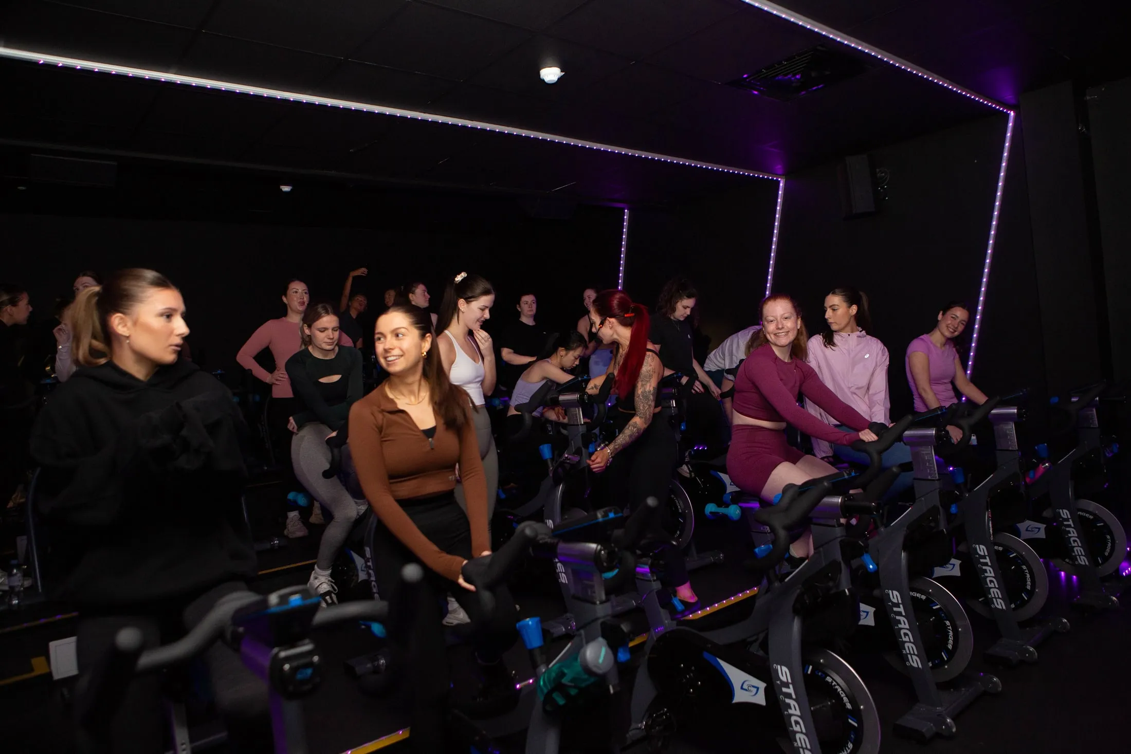 Women smiling and chatting at spin bikes in a dark studio edged with purple lights