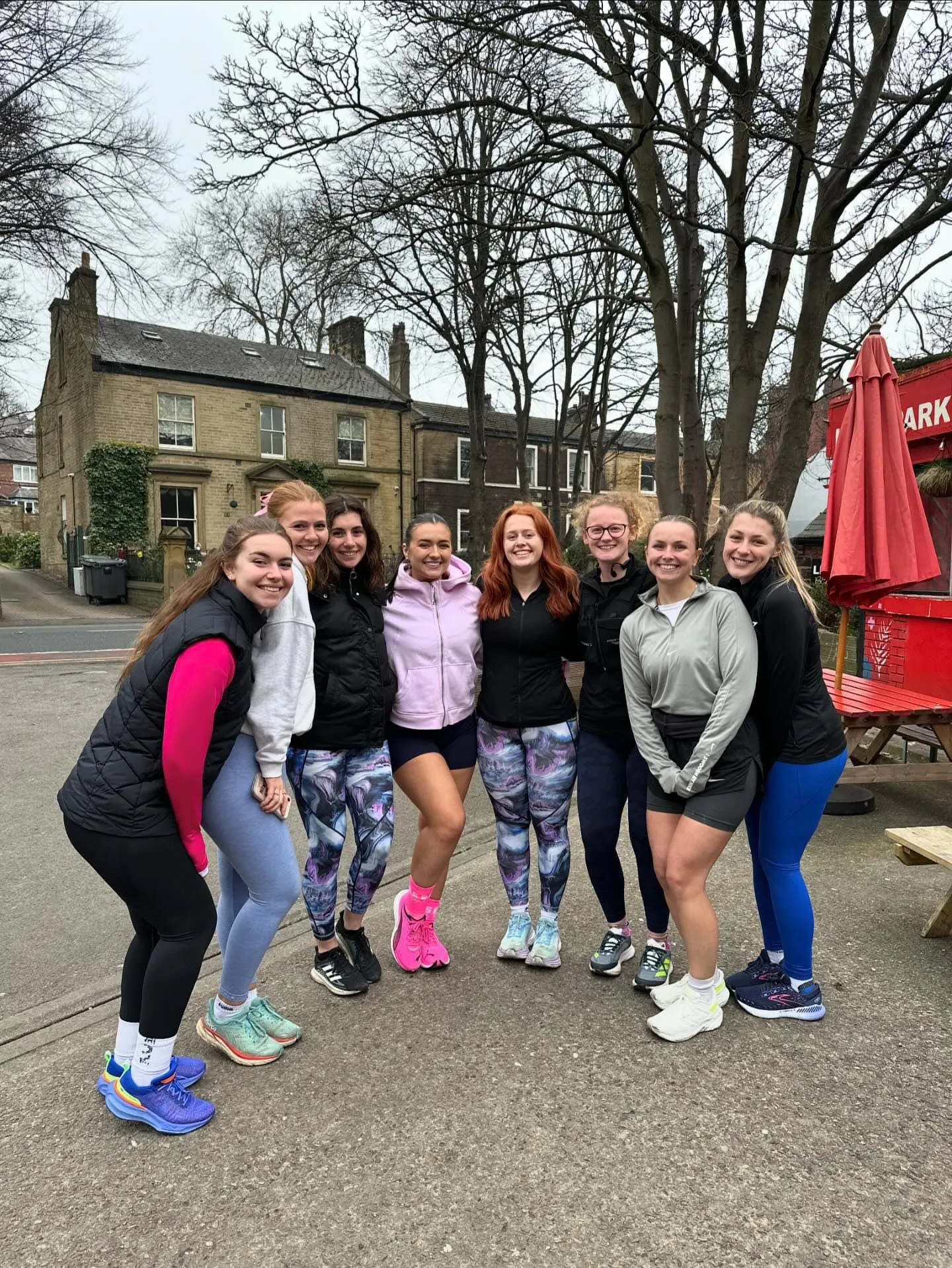 Eight women in activewear posing together on a street, with bare trees and stone houses behind
