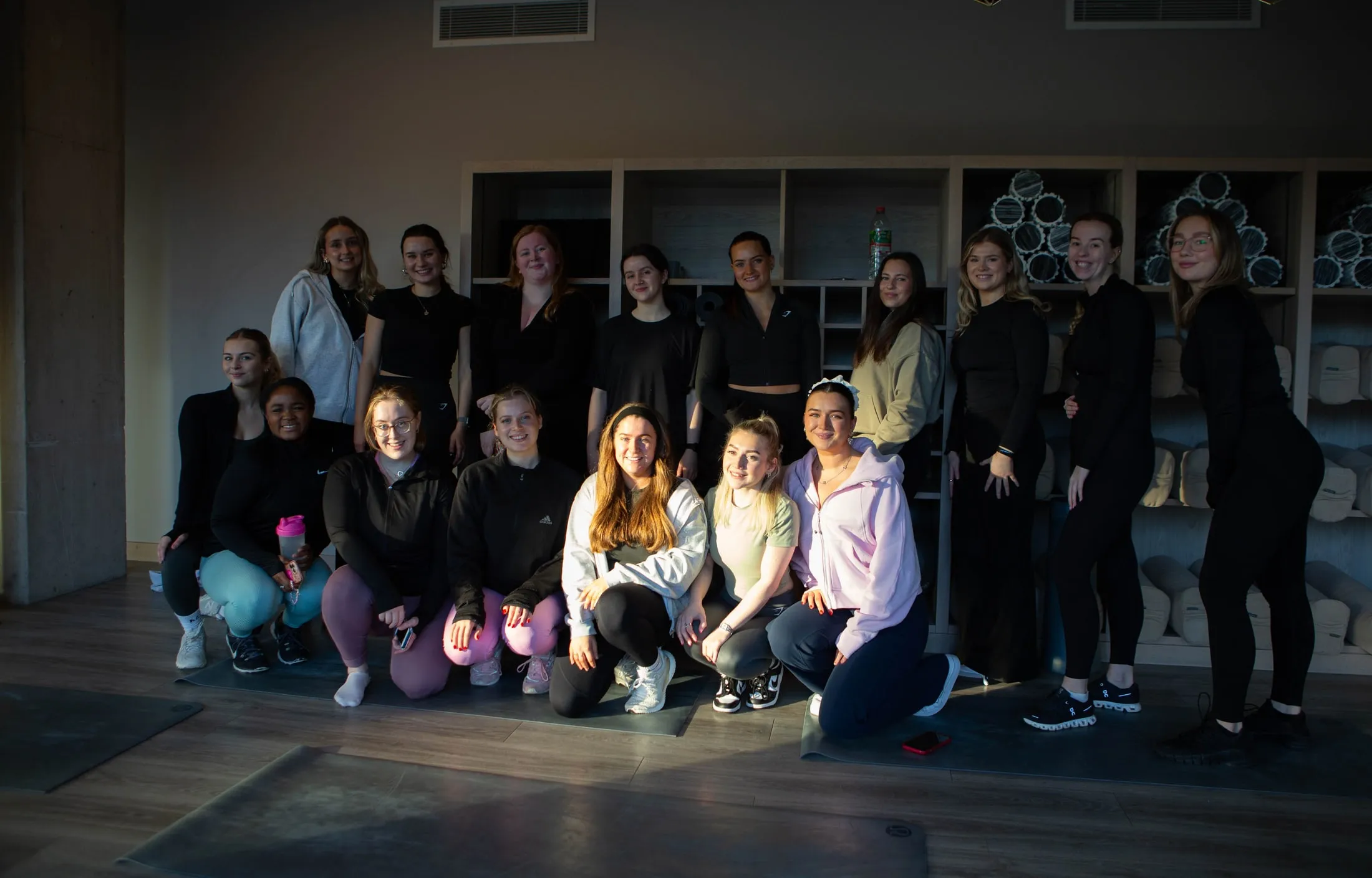 A group of women posing together in a studio, in front of shelves of rolled-up mats