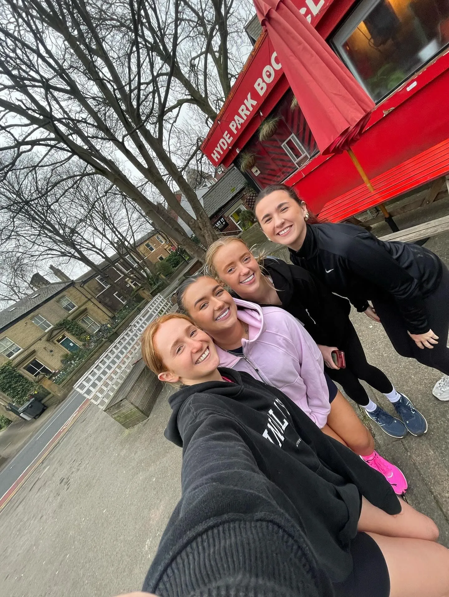 A tilted selfie of four smiling women in activewear outside a red building under bare trees