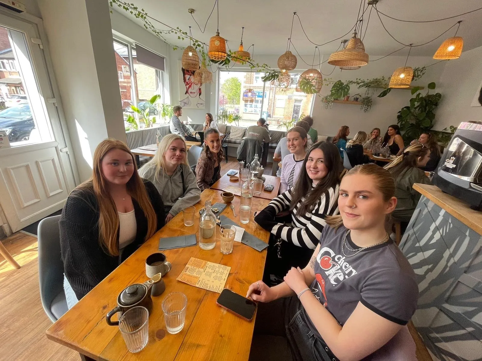 Six women round a table in a bright café with wicker lampshades and trailing plants