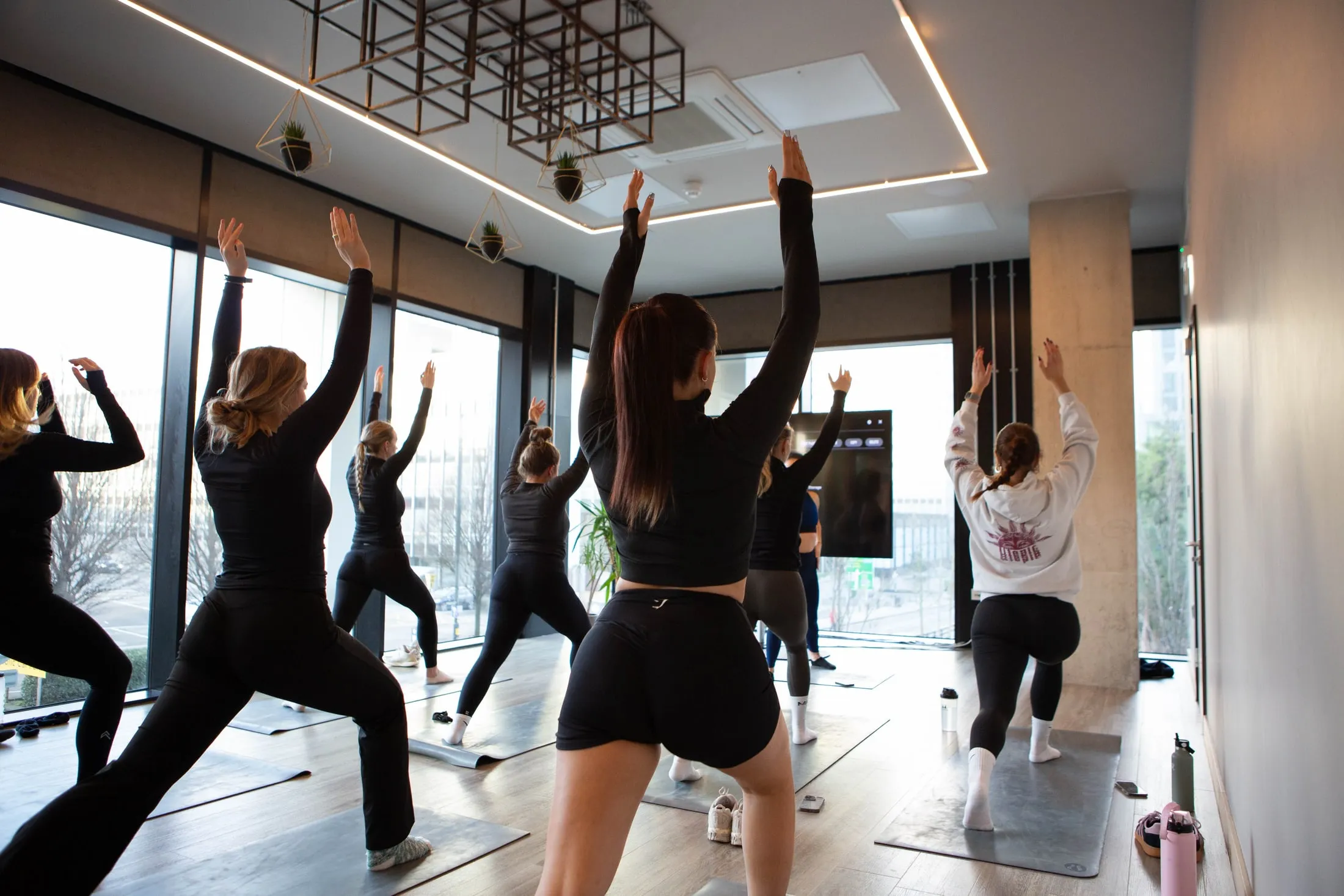 Women lunging with arms raised on yoga mats by tall windows, seen from behind