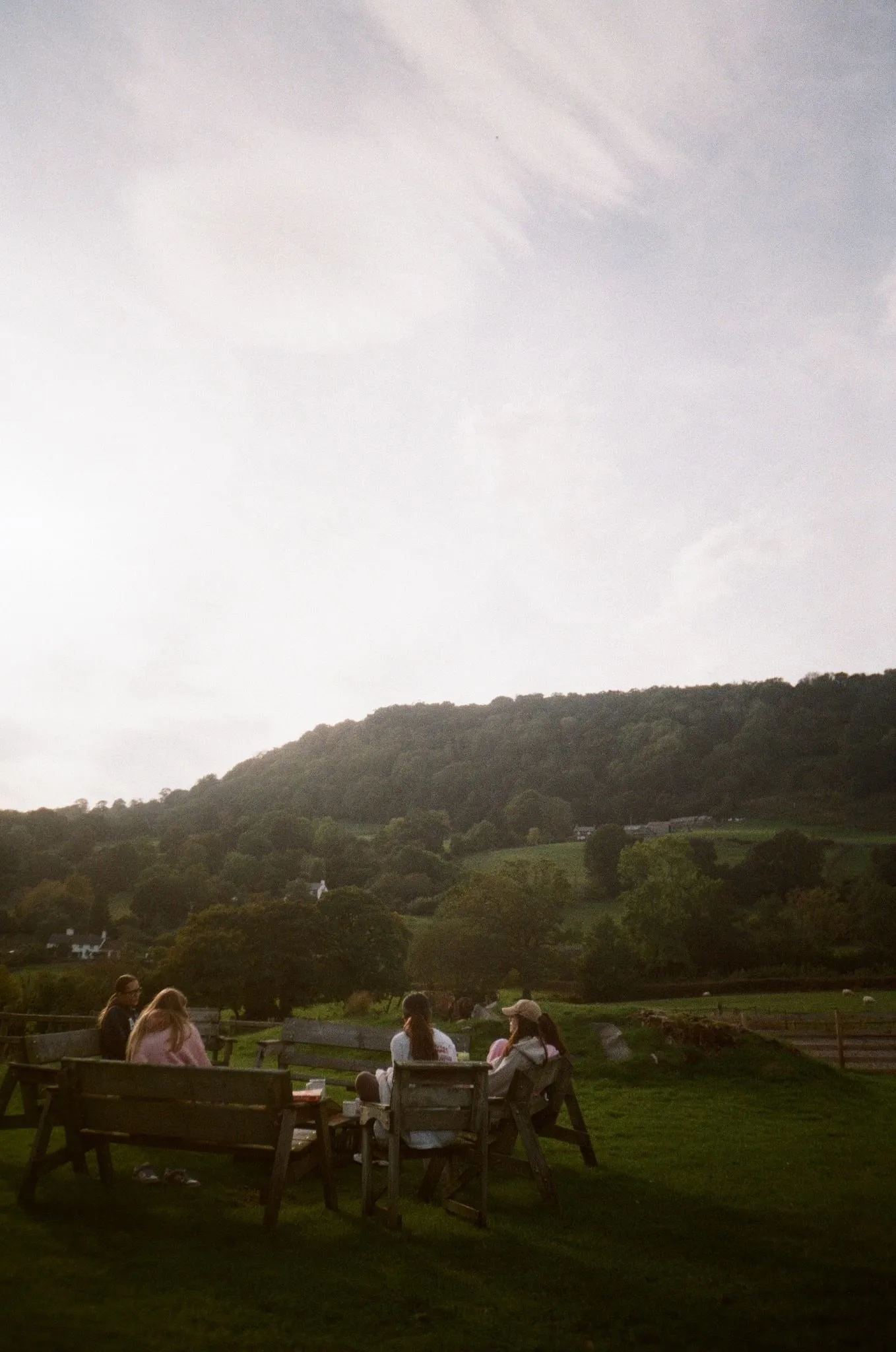 A small group of women on wooden benches on a lawn below a wooded hill, shot on film