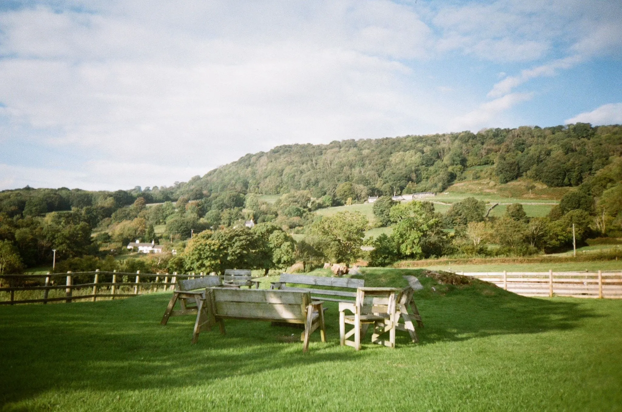 Wooden benches in a circle on a sunny lawn, wooded hills and fields beyond, shot on film