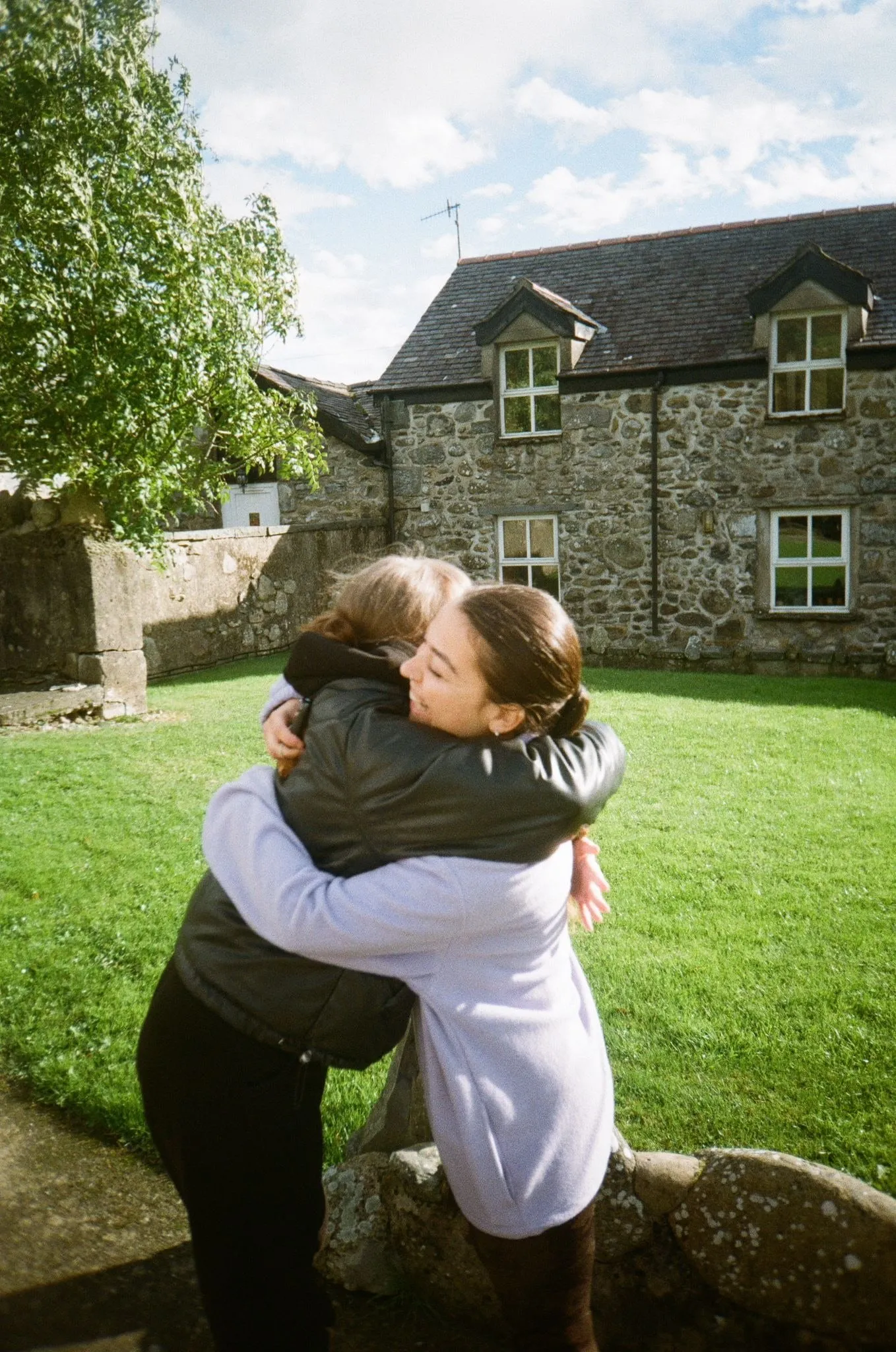 Two women hugging on a lawn in front of a stone cottage, shot on film