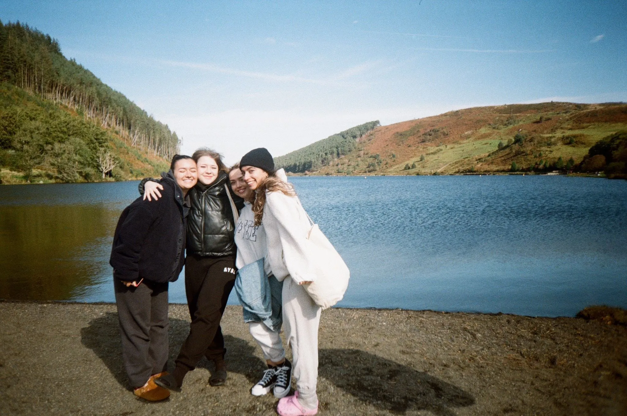 Four women hugging on a lakeshore with hills behind, shot on film