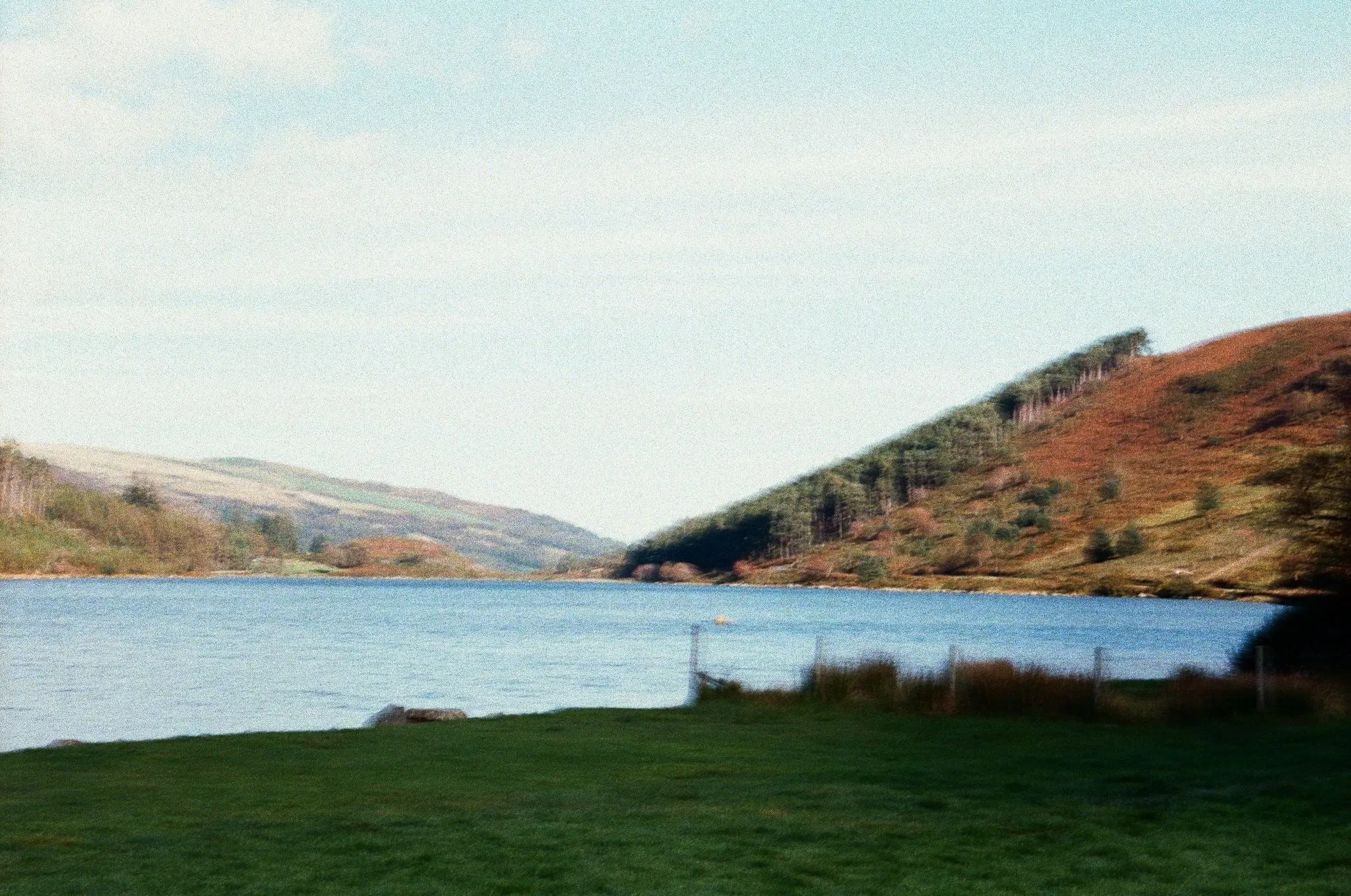 A blue lake between russet hills, a grassy bank and fence in front, shot on film