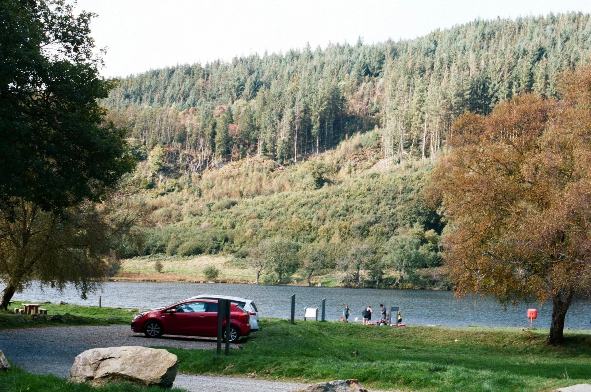 Cars parked by a lake below steep forested hills, people at the water’s edge, shot on film