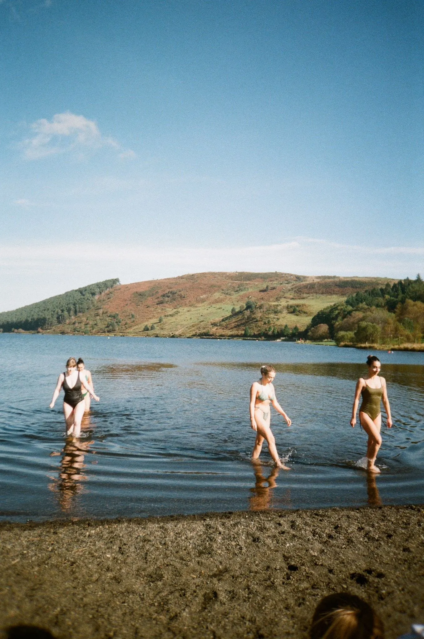Three women wading into a lake in swimsuits, green hills behind, shot on film