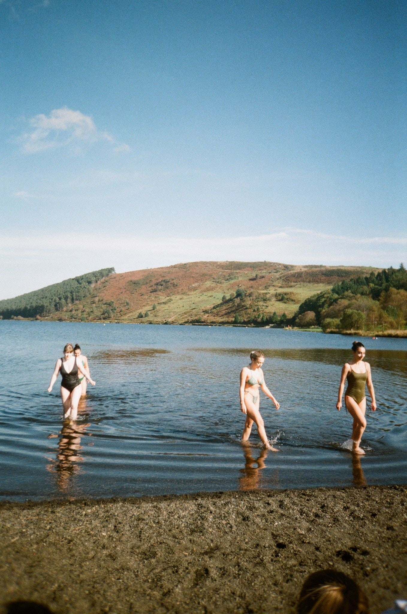Three women wading into a lake in swimsuits, green hills behind, shot on film