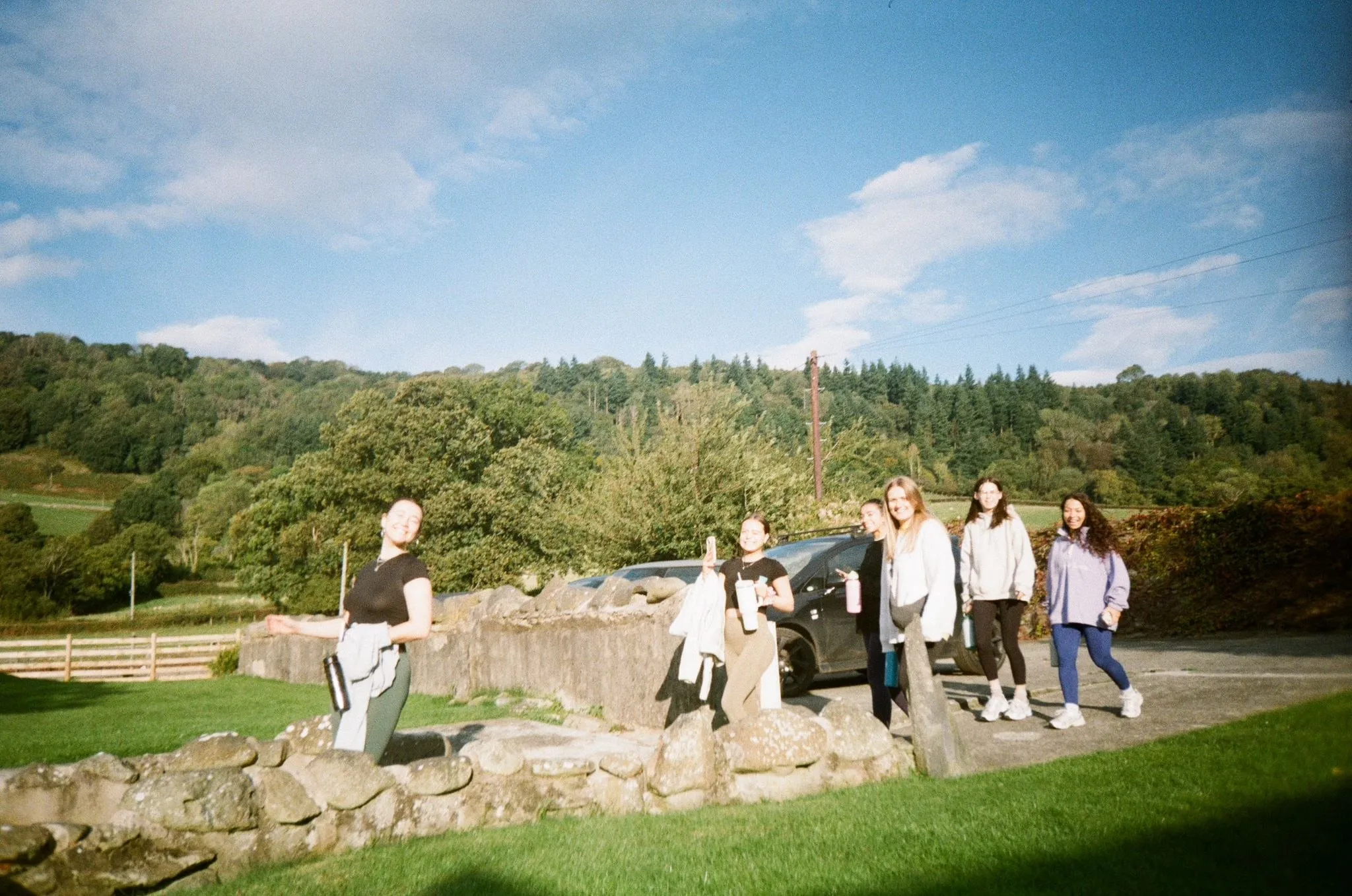 Women with water bottles walking past a low stone wall below wooded hills, shot on film