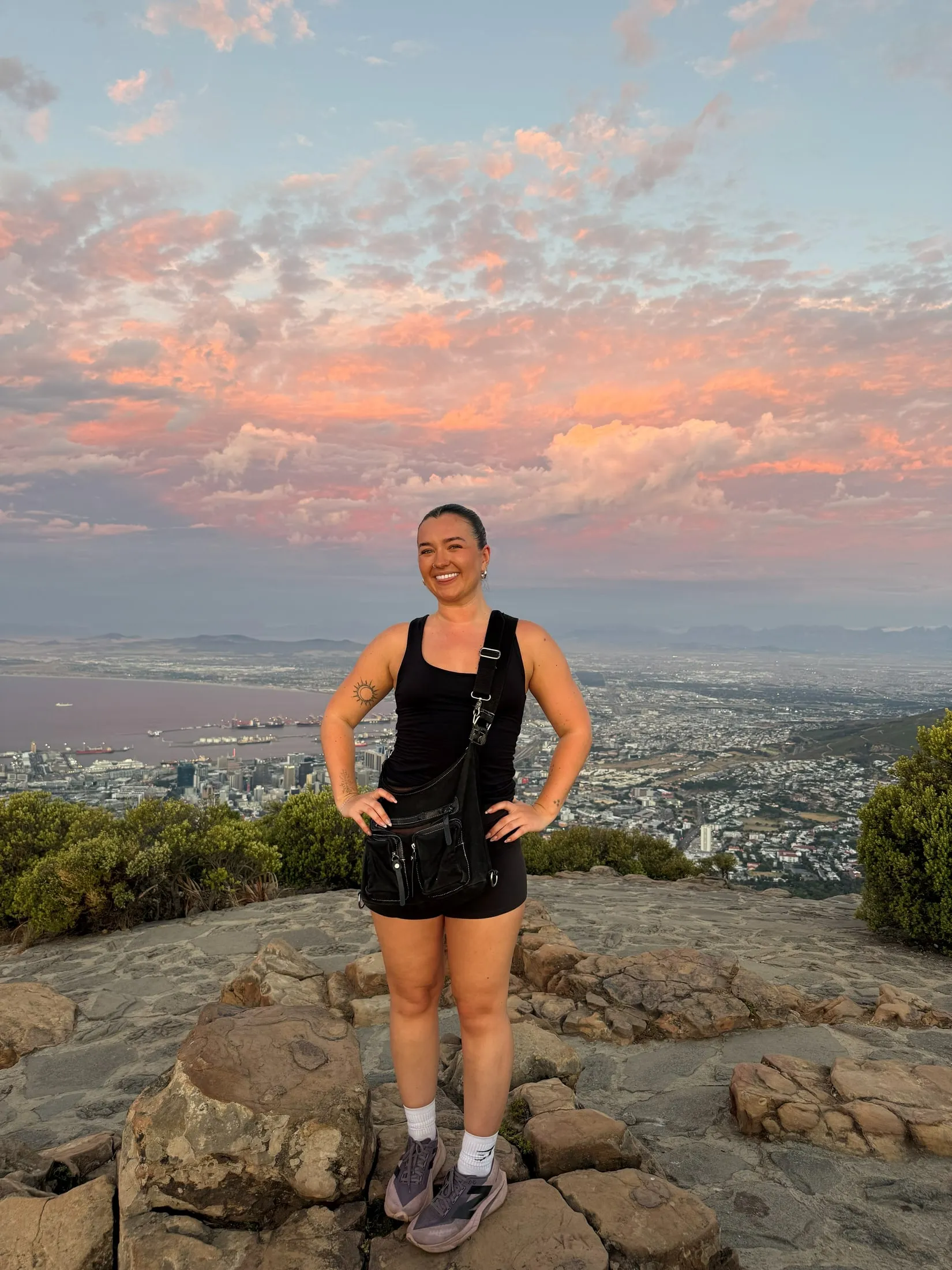 Megan standing on rocks at sunset, with a pink sky and a city far below