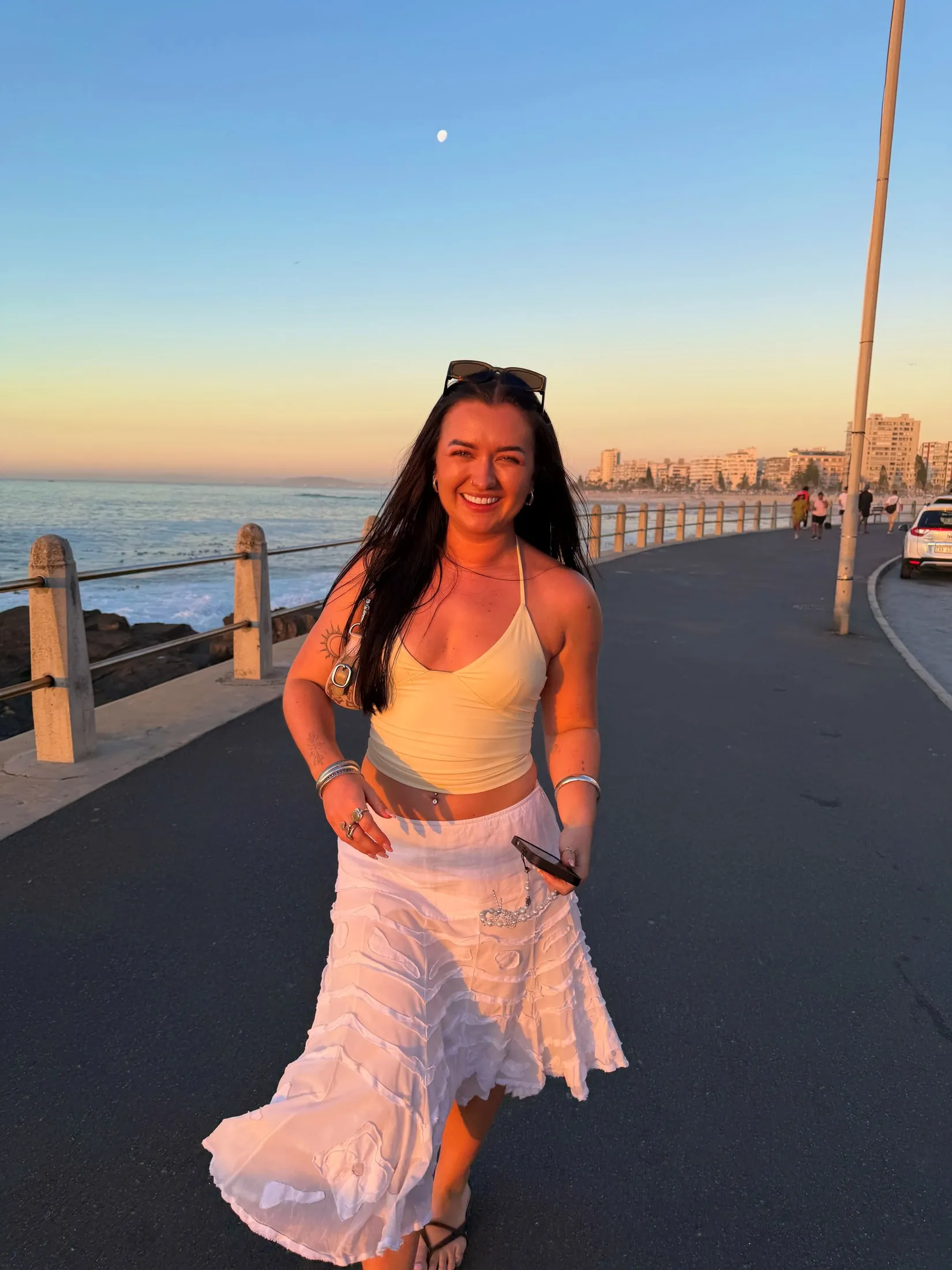 Megan walking along a seafront at sunset in a white skirt