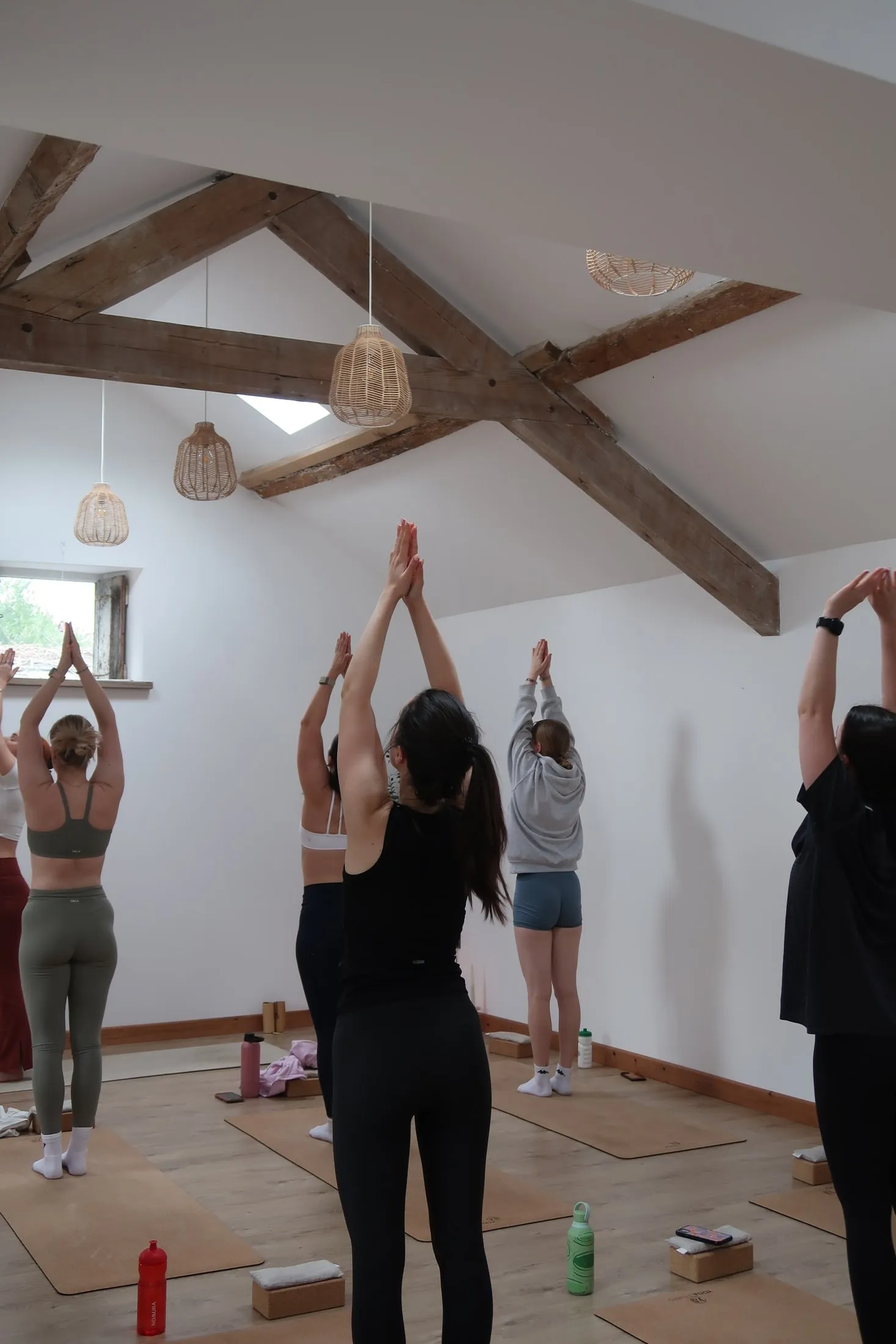 Women on yoga mats reaching their arms overhead, palms together, in a white beamed studio