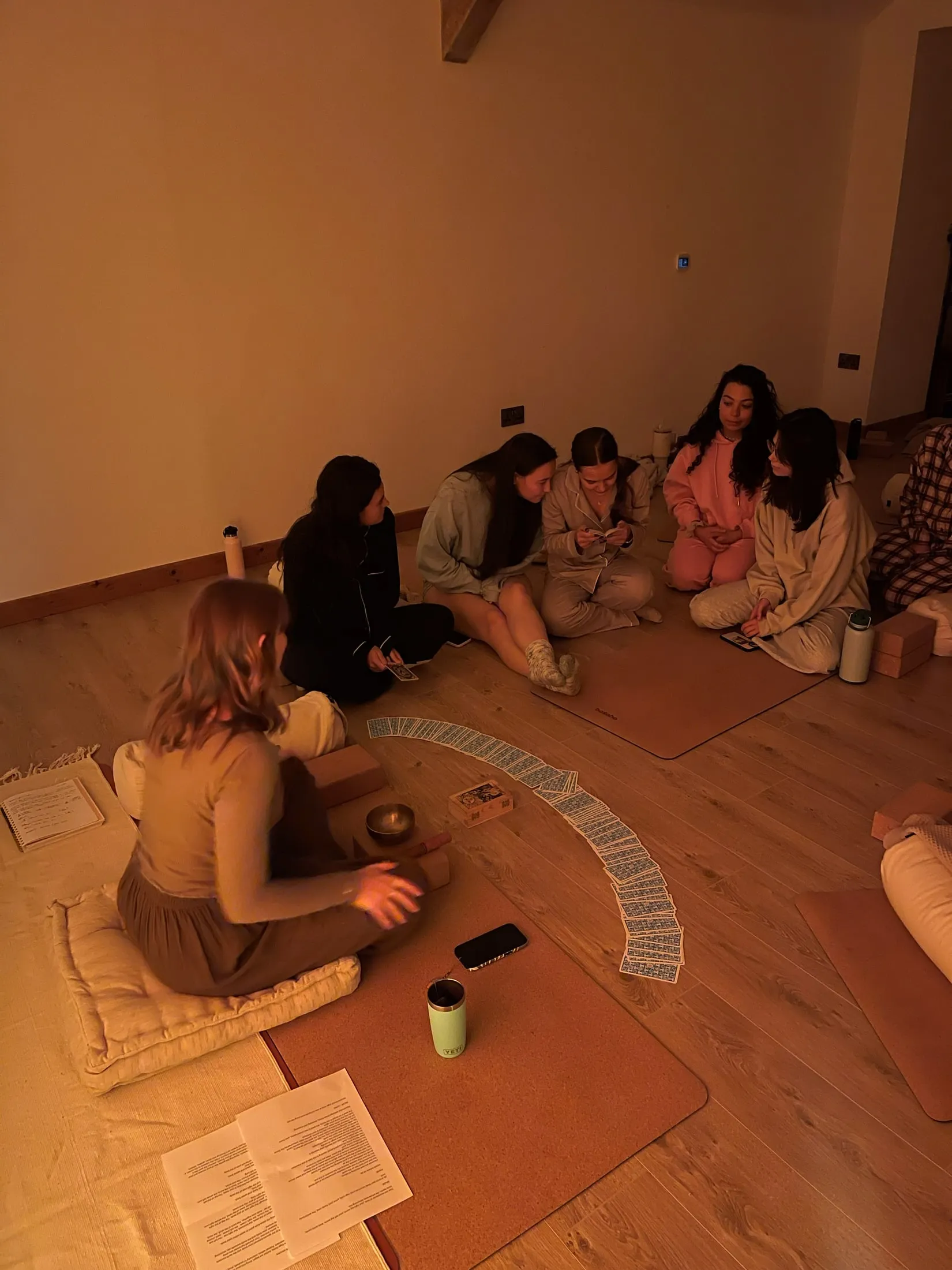 Women sitting in a circle on the floor in warm low light, a long arc of cards laid out between them