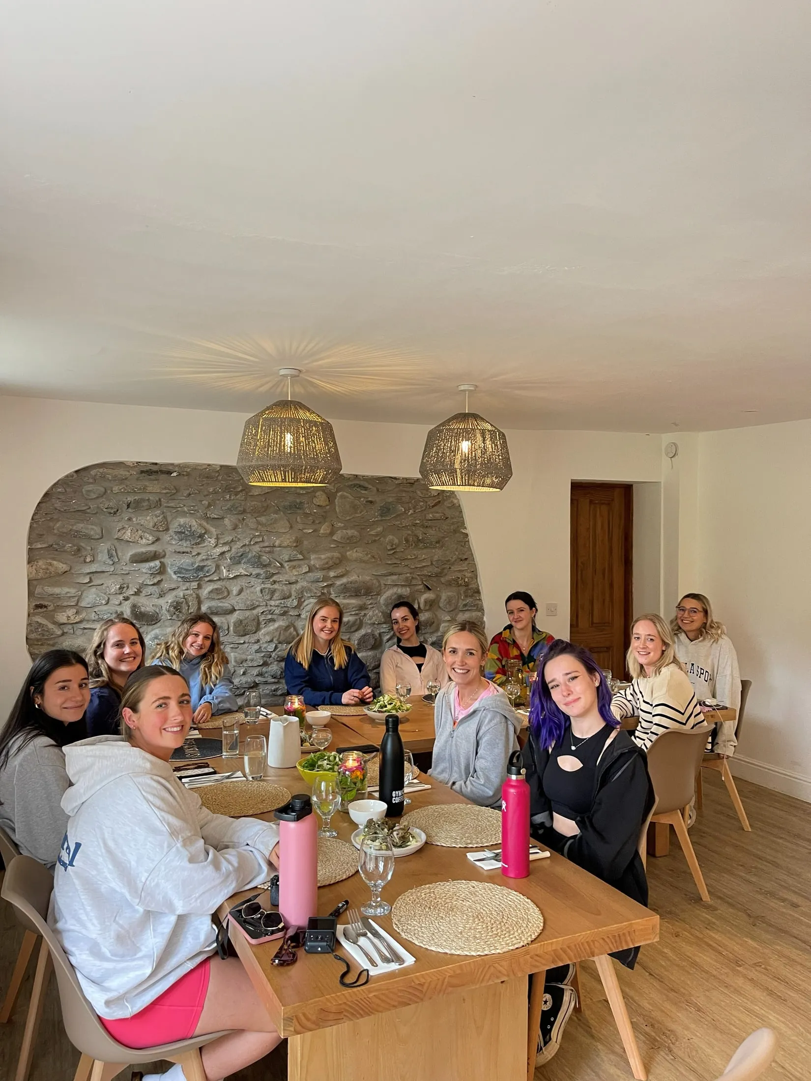 A group of women smiling round a long dining table, woven lamps above and a stone wall behind